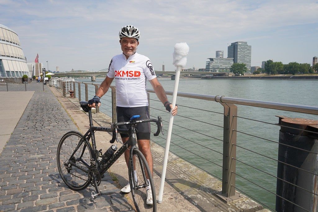 Helmut Thillmann mit einem Fahrrad und einem großen Wattestäbchen. Im Hintergrund ist ein Geländer, dahinter ist ein Gewässer. Helmut Thillmann mit einem Fahrrad und einem großen Wattestäbchen. Im Hintergrund ist ein Geländer, dahinter ist ein Gewässer.