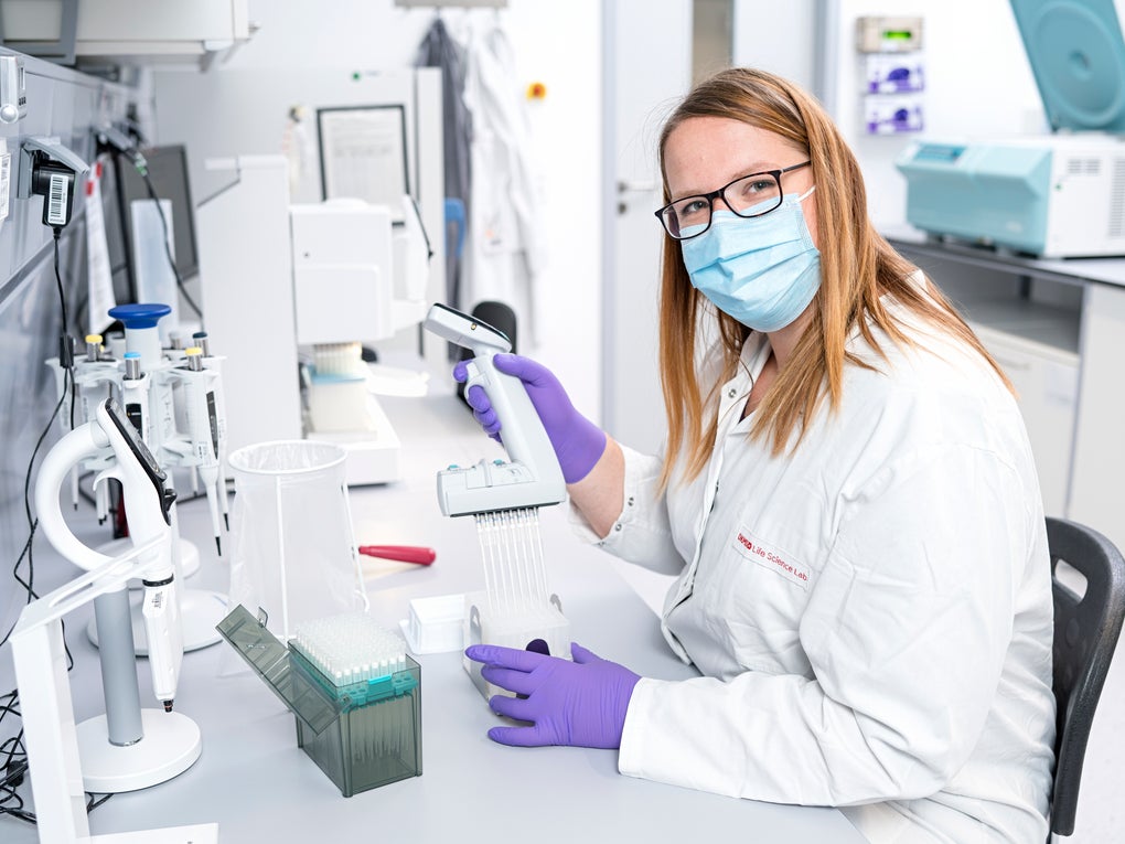 Christin Paech trägt Maske und Handschuhe in einem Labor. Sie hält ein Objekt in den Händen Christin Paech trägt Maske und Handschuhe in einem Labor. Sie hält ein Objekt in den Händen