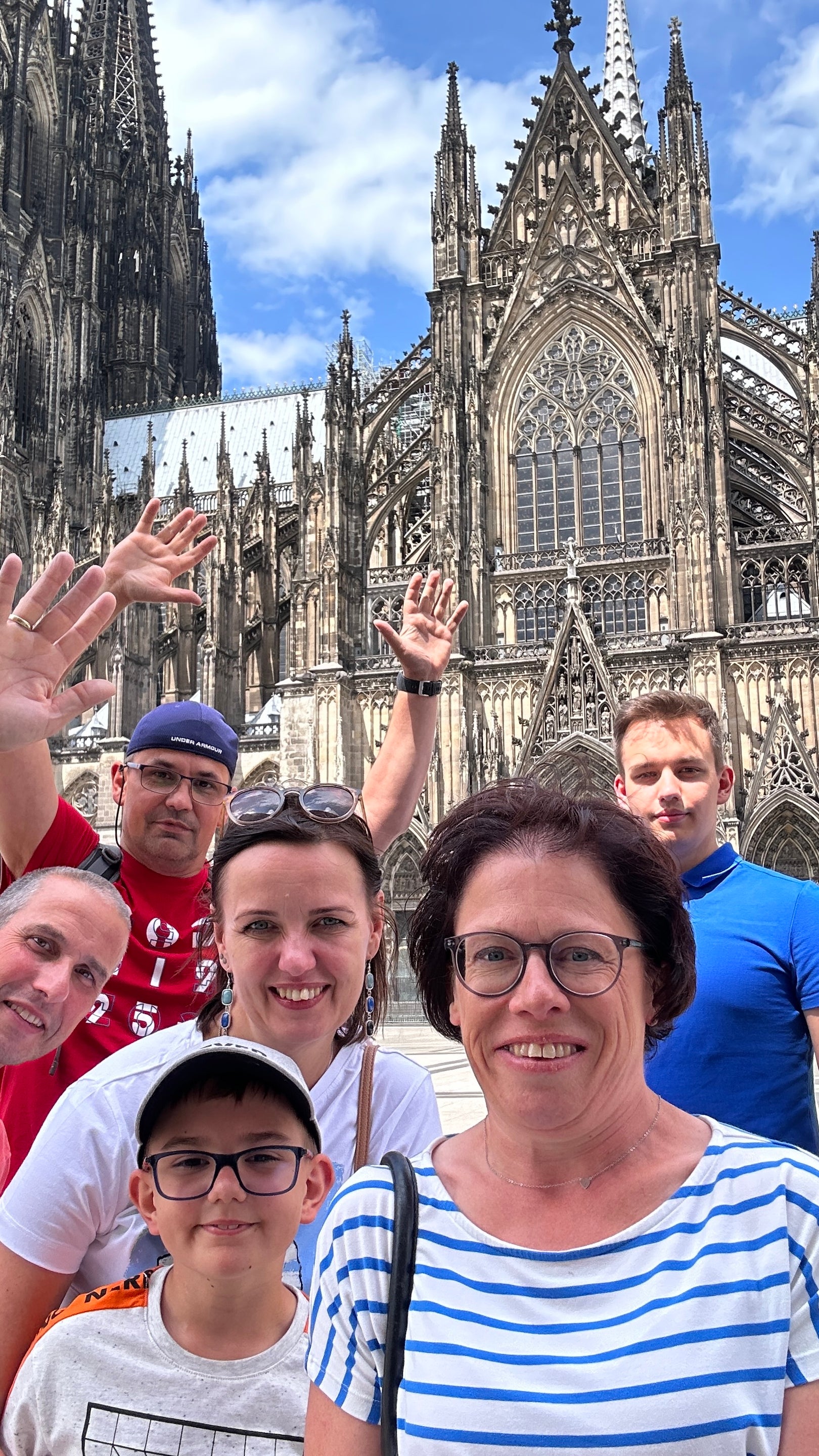 Sieben Personen stehen eng zusammen vor dem Kölner Dom und blicken in die Kamera. Einige winken, andere lächeln. Im Vordergrund stehen zwei Frauen und ein Junge mit Brille, im Hintergrund sind Erwachsene in farbigen T-Shirts zu sehen. Der Kölner Dom mit seinen markanten Türmen und Fassadendetails füllt den Hintergrund. Sieben Personen stehen eng zusammen vor dem Kölner Dom und blicken in die Kamera. Einige winken, andere lächeln. Im Vordergrund stehen zwei Frauen und ein Junge mit Brille, im Hintergrund sind Erwachsene in farbigen T-Shirts zu sehen. Der Kölner Dom mit seinen markanten Türmen und Fassadendetails füllt den Hintergrund.