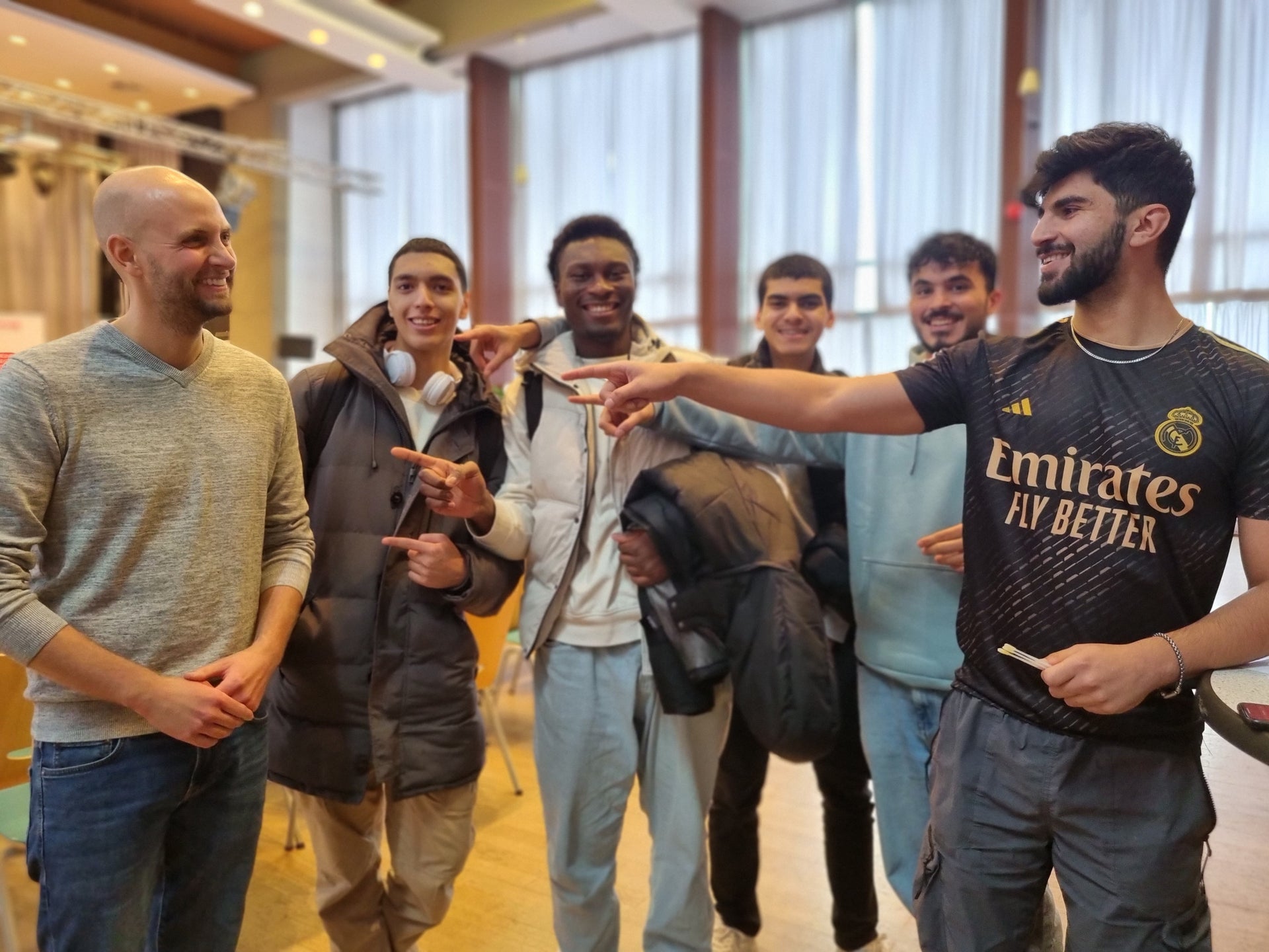 Sechs junge Männer stehen in einem Innenraum mit hohen Fenstern nebeneinander. Fünf von ihnen zeigen lachend auf den Mann ganz links, der ein graues Oberteil trägt und ebenfalls lächelt. Einige der Personen tragen Jacken oder halten sie in der Hand. Der Raum ist hell beleuchtet, im Hintergrund sind Vorhänge und Scheinwerfer zu sehen. Sechs junge Männer stehen in einem Innenraum mit hohen Fenstern nebeneinander. Fünf von ihnen zeigen lachend auf den Mann ganz links, der ein graues Oberteil trägt und ebenfalls lächelt. Einige der Personen tragen Jacken oder halten sie in der Hand. Der Raum ist hell beleuchtet, im Hintergrund sind Vorhänge und Scheinwerfer zu sehen.