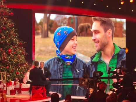 Eine Bühne, auf der ein großer Weihnachtsbaum steht. Im Hintergrund ein Bildschirm, auf der man einen Mann und einen Jungen sieht Eine Bühne, auf der ein großer Weihnachtsbaum steht. Im Hintergrund ein Bildschirm, auf der man einen Mann und einen Jungen sieht