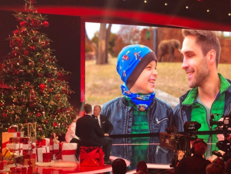 Eine Bühne, auf der ein großer Weihnachtsbaum steht. Im Hintergrund ein Bildschirm, auf der man einen Mann und einen Jungen sieht Eine Bühne, auf der ein großer Weihnachtsbaum steht. Im Hintergrund ein Bildschirm, auf der man einen Mann und einen Jungen sieht