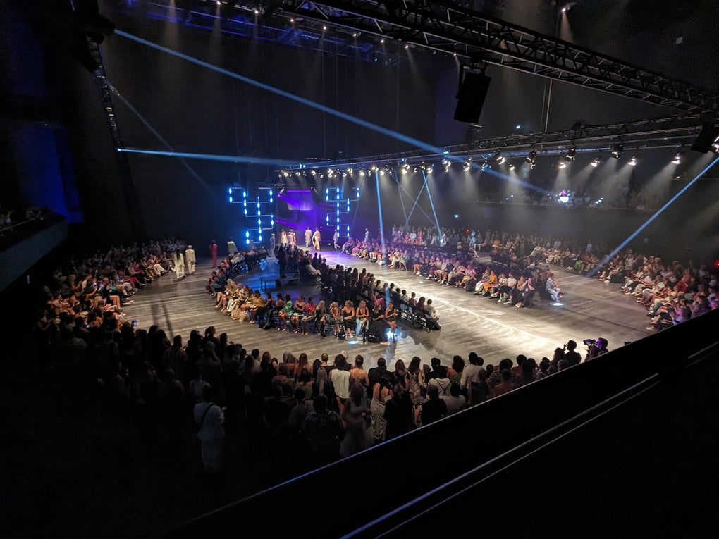 Blick in eine große Veranstaltungshalle mit kreisförmigem Laufsteg. Rundherum sitzen zahlreiche Zuschauerinnen und Zuschauer. Die Bühne ist mit Lichtinstallationen und Spots ausgeleuchtet, in der Mitte steht eine Gruppe von Menschen. Blick in eine große Veranstaltungshalle mit kreisförmigem Laufsteg. Rundherum sitzen zahlreiche Zuschauerinnen und Zuschauer. Die Bühne ist mit Lichtinstallationen und Spots ausgeleuchtet, in der Mitte steht eine Gruppe von Menschen.