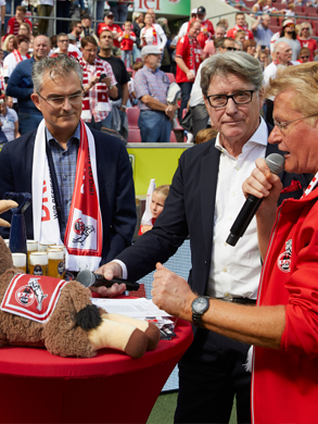 Eine Gruppe von Männern steht um einen Tisch mit einem Ziegen Kuscheltier