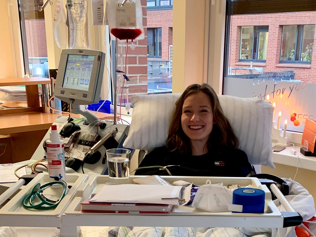 Eine junge Frau liegt lächelnd in einem Behandlungsstuhl während einer peripheren Stammzellentnahme. Vor ihr steht eine Apheresemaschine mit einem Infusionsbeutel, in dem sich Blutbestandteile befinden. Auf dem Rolltisch vor ihr liegen medizinische Utensilien und ein Glas Wasser. Im Hintergrund ist ein Fenster mit Blick auf ein Gebäude aus rotem Backstein sowie eine Kerze und eine Lichterkette auf der Fensterbank zu sehen. Eine junge Frau liegt lächelnd in einem Behandlungsstuhl während einer peripheren Stammzellentnahme. Vor ihr steht eine Apheresemaschine mit einem Infusionsbeutel, in dem sich Blutbestandteile befinden. Auf dem Rolltisch vor ihr liegen medizinische Utensilien und ein Glas Wasser. Im Hintergrund ist ein Fenster mit Blick auf ein Gebäude aus rotem Backstein sowie eine Kerze und eine Lichterkette auf der Fensterbank zu sehen.