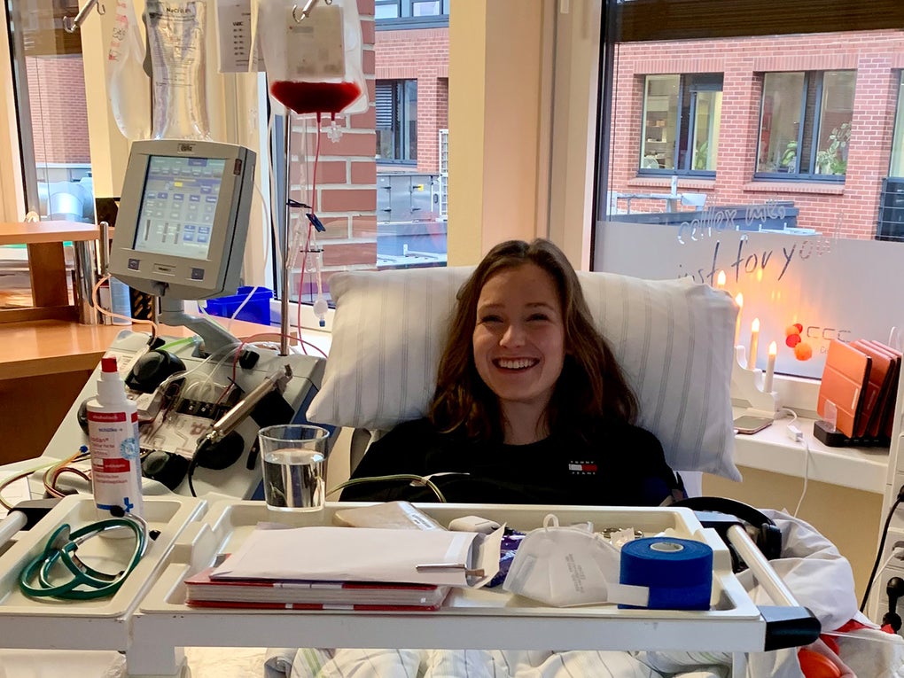 Eine junge Frau liegt lächelnd in einem Behandlungsstuhl während einer peripheren Stammzellentnahme. Vor ihr steht eine Apheresemaschine mit einem Infusionsbeutel, in dem sich Blutbestandteile befinden. Auf dem Rolltisch vor ihr liegen medizinische Utensilien und ein Glas Wasser. Im Hintergrund ist ein Fenster mit Blick auf ein Gebäude aus rotem Backstein sowie eine Kerze und eine Lichterkette auf der Fensterbank zu sehen. Eine junge Frau liegt lächelnd in einem Behandlungsstuhl während einer peripheren Stammzellentnahme. Vor ihr steht eine Apheresemaschine mit einem Infusionsbeutel, in dem sich Blutbestandteile befinden. Auf dem Rolltisch vor ihr liegen medizinische Utensilien und ein Glas Wasser. Im Hintergrund ist ein Fenster mit Blick auf ein Gebäude aus rotem Backstein sowie eine Kerze und eine Lichterkette auf der Fensterbank zu sehen.