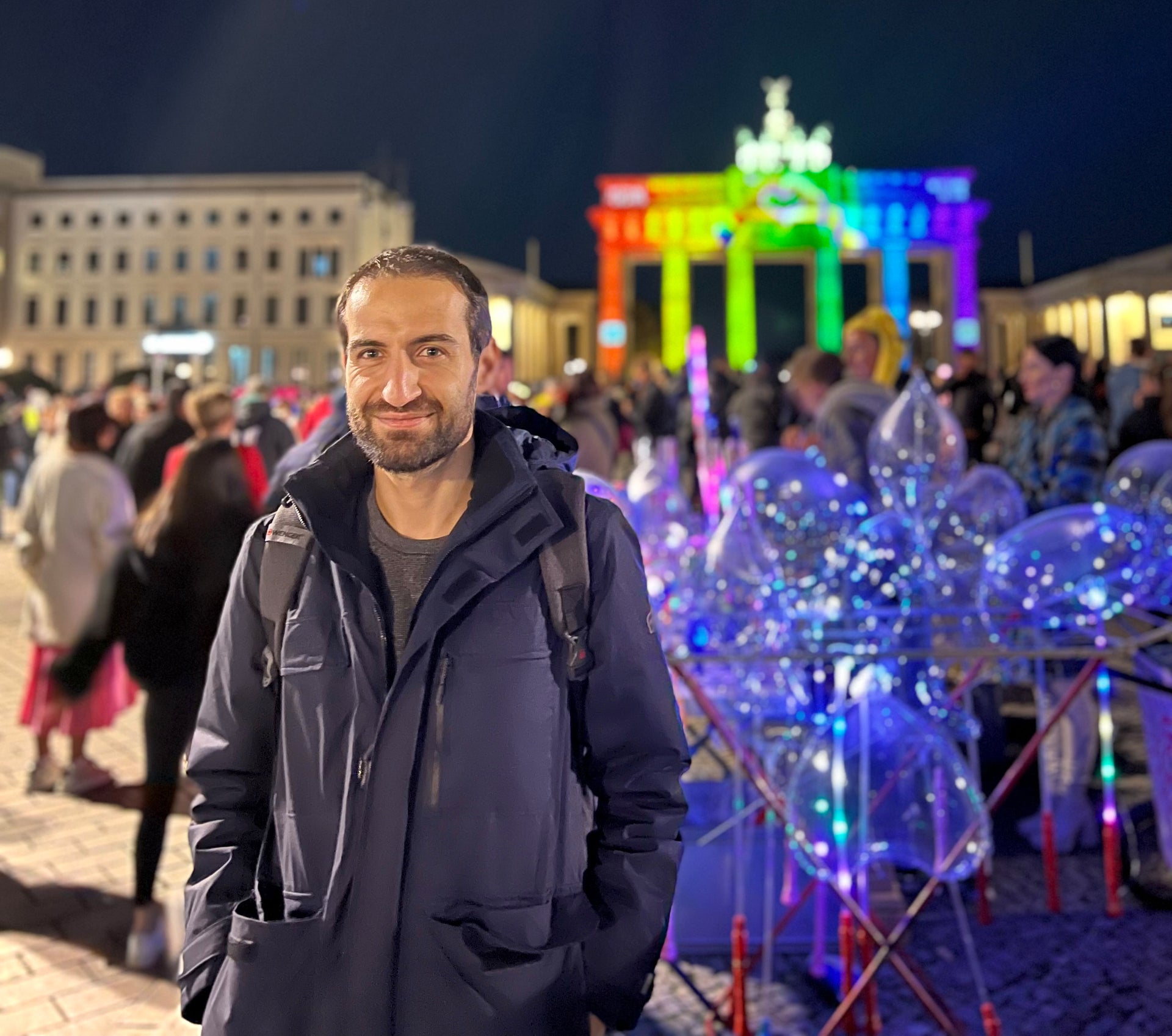 Ein Mann steht abends auf einem belebten Platz vor dem Brandenburger Tor in Berlin, das in Regenbogenfarben beleuchtet ist. Er trägt eine dunkelblaue Jacke und blickt lächelnd in die Kamera. Um ihn herum sind weitere Personen und beleuchtete transparente Luftballons zu sehen. Ein Mann steht abends auf einem belebten Platz vor dem Brandenburger Tor in Berlin, das in Regenbogenfarben beleuchtet ist. Er trägt eine dunkelblaue Jacke und blickt lächelnd in die Kamera. Um ihn herum sind weitere Personen und beleuchtete transparente Luftballons zu sehen.