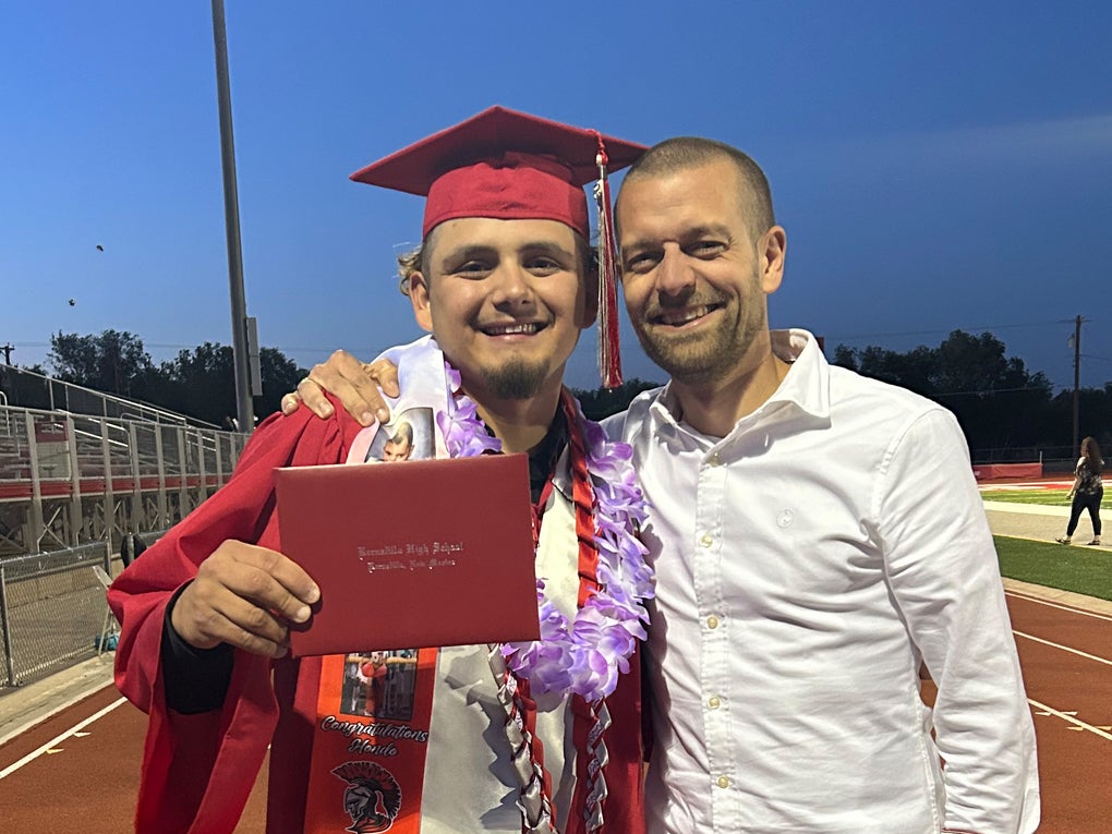 Zwei junge Männer lachen am Rande eine Sportfelds in die Kamera. Der eine trägt eine typische Robe zum Highschool-Abschluss. Zwei junge Männer lachen am Rande eine Sportfelds in die Kamera. Der eine trägt eine typische Robe zum Highschool-Abschluss.