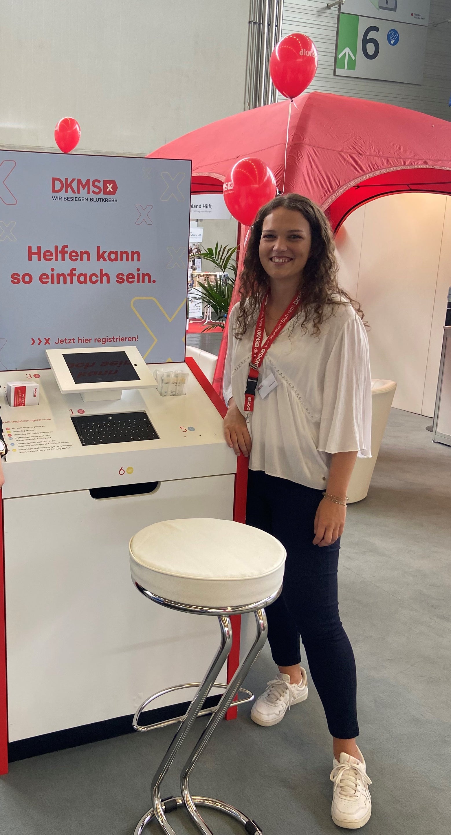 Eine Frau steht an einem DKMS-Informationsstand in einer Messehalle. Sie trägt ein weißes Oberteil, eine dunkelblaue Hose und weiße Turnschuhe. Um ihren Hals hängt ein Namensschild an einem roten Band. Auf dem Bildschirm am Stand steht „Helfen kann so einfach sein“. Im Hintergrund sind ein rotes Zelt, rote Luftballons und ein Schild mit der Hallenkennzeichnung „6“ zu sehen. Eine Frau steht an einem DKMS-Informationsstand in einer Messehalle. Sie trägt ein weißes Oberteil, eine dunkelblaue Hose und weiße Turnschuhe. Um ihren Hals hängt ein Namensschild an einem roten Band. Auf dem Bildschirm am Stand steht „Helfen kann so einfach sein“. Im Hintergrund sind ein rotes Zelt, rote Luftballons und ein Schild mit der Hallenkennzeichnung „6“ zu sehen.