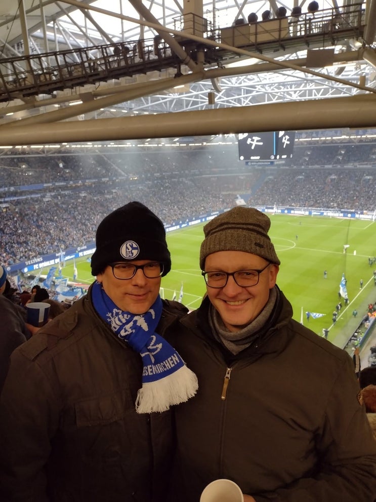 Patient Martin (l.) ist gemeinsam mit seinem Freund Michael (r.) bei einem FC Schalke 04 Spiel. 
Um seinen Hals trägt Martin einen blau-weißen FC Schalke Schal und eine Mütze. 