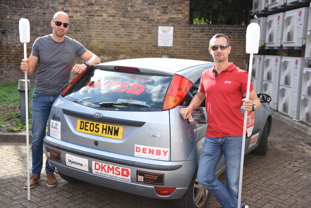 Zwei Männer stehen neben einem Auto und halten große Wattestäbchen Zwei Männer stehen neben einem Auto und halten große Wattestäbchen