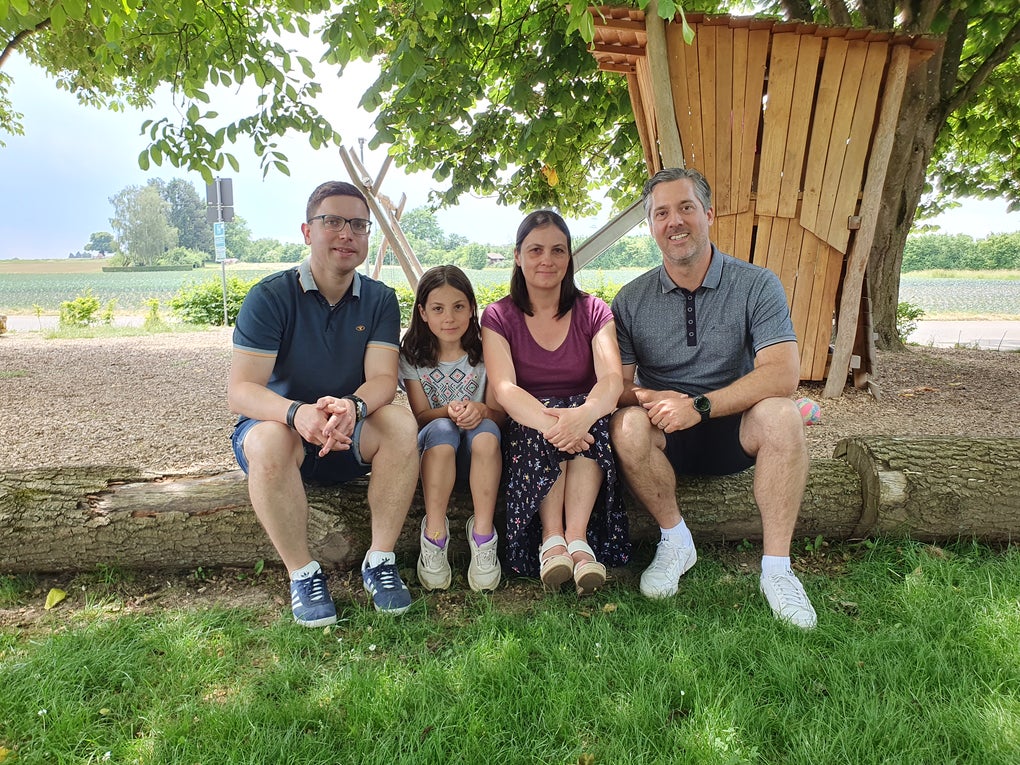 Vier Personen sitzen auf einem Baumstamm im Schatten eines Baumes. Zwei Erwachsene und ein Kind sitzen in der Mitte, ein weiterer Erwachsener sitzt rechts außen. Im Hintergrund sind ein Baumhaus aus Holz, ein Feldweg und grüne Felder zu sehen. Alle Personen blicken in die Kamera.
Vier Personen sitzen auf einem Baumstamm im Schatten eines Baumes. Zwei Erwachsene und ein Kind sitzen in der Mitte, ein weiterer Erwachsener sitzt rechts außen. Im Hintergrund sind ein Baumhaus aus Holz, ein Feldweg und grüne Felder zu sehen. Alle Personen blicken in die Kamera.