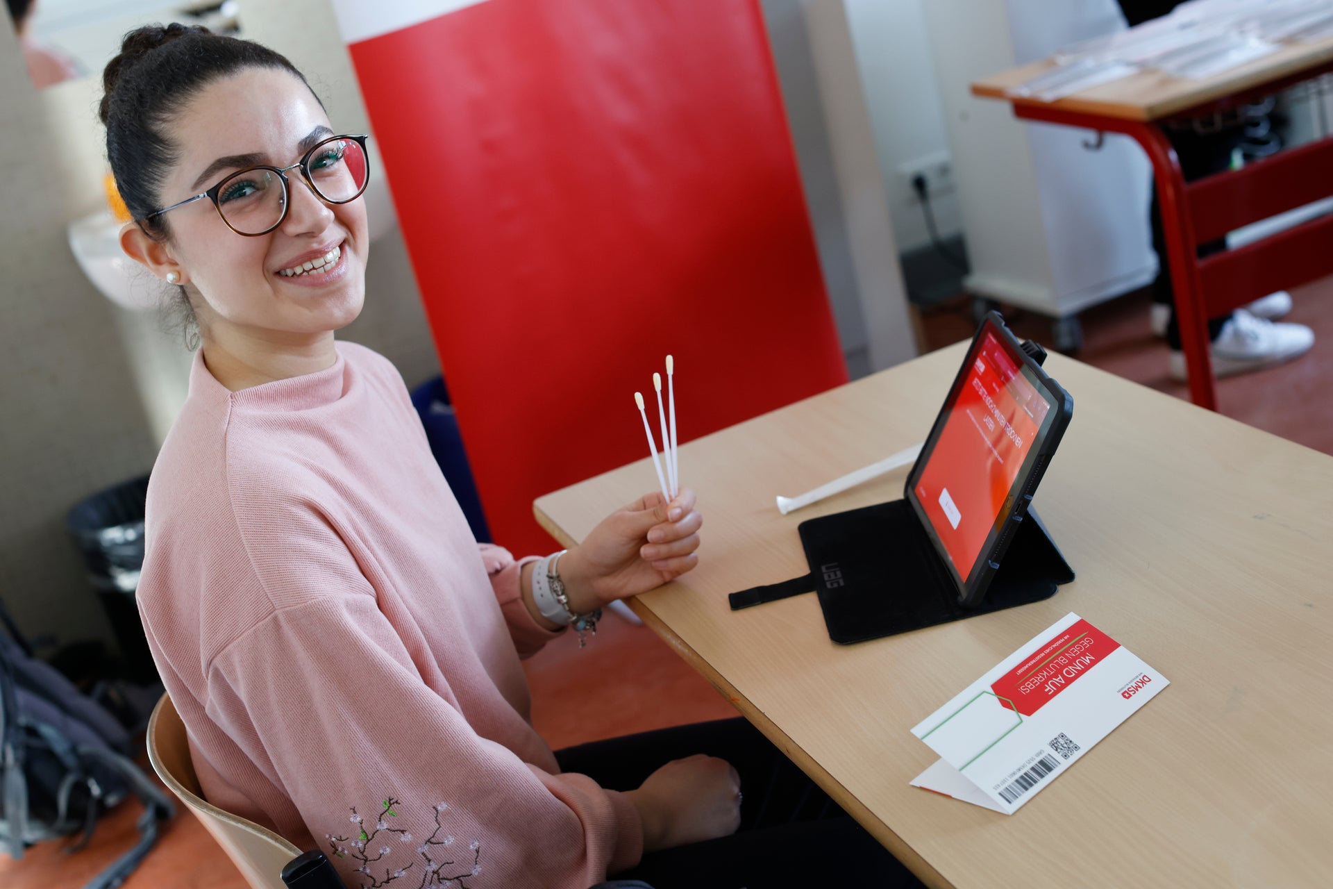 Eine Frau sitzt an einem Tisch mit einem Tablet und hält drei Wattestäbchen zur Registrierung als Stammzellspenderin in der Hand Eine Frau sitzt an einem Tisch mit einem Tablet und hält drei Wattestäbchen zur Registrierung als Stammzellspenderin in der Hand