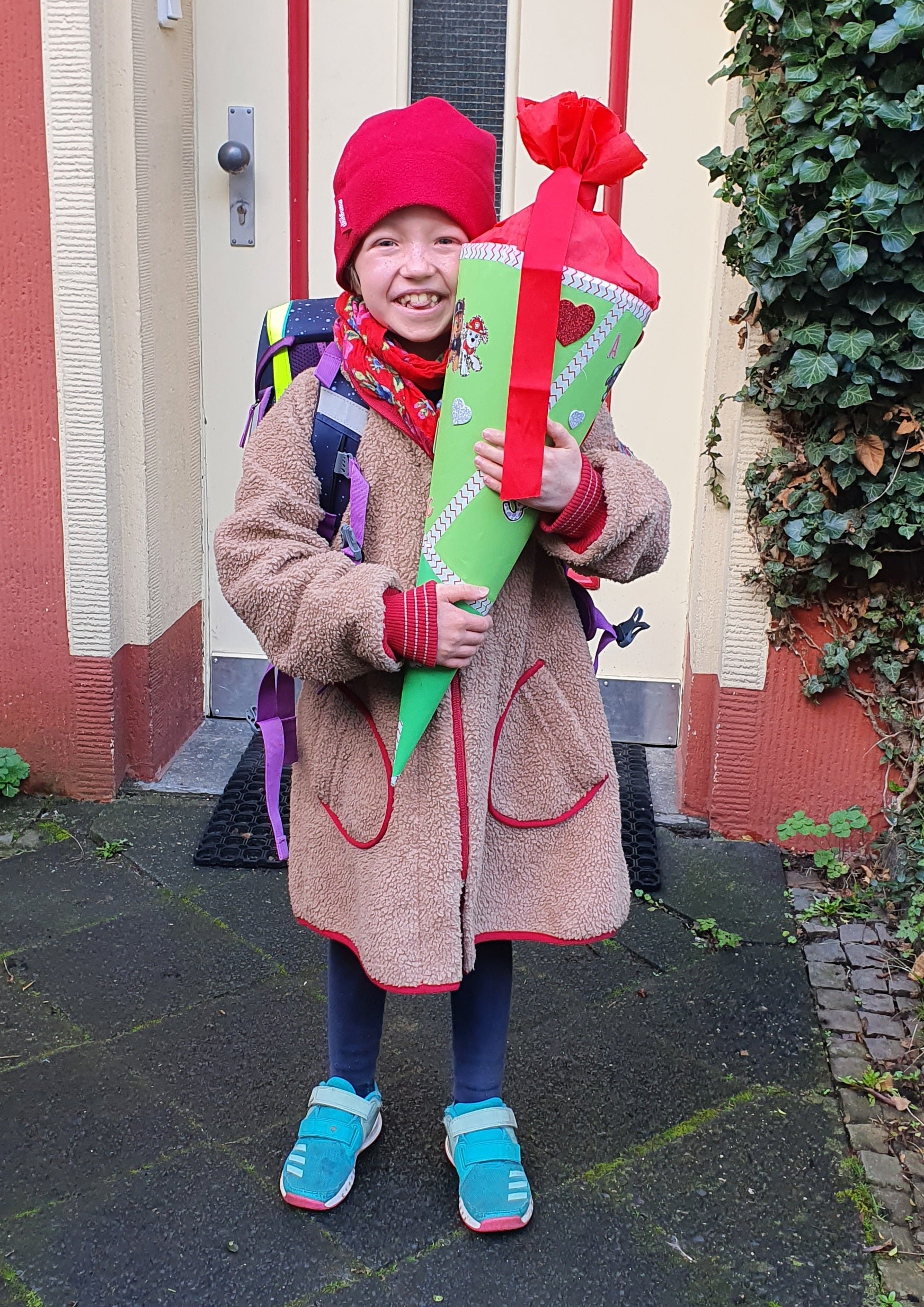 Ein fröhliches Kind steht vor einer Haustür und hält stolz eine große, grün-rote Schultüte im Arm. Es trägt eine rote Mütze, eine beigefarbene Jacke mit roten Nähten, blaue Leggings und türkisfarbene Schuhe. Auf dem Rücken ist ein Schulranzen zu sehen. Das Kind lächelt breit in die Kamera. Es sieht nach einem besonderen Tag aus, vermutlich ein erster Schultag oder Schulanfang nach einer Pause. Rechts im Bild wächst Efeu an der Hauswand.
Ein fröhliches Kind steht vor einer Haustür und hält stolz eine große, grün-rote Schultüte im Arm. Es trägt eine rote Mütze, eine beigefarbene Jacke mit roten Nähten, blaue Leggings und türkisfarbene Schuhe. Auf dem Rücken ist ein Schulranzen zu sehen. Das Kind lächelt breit in die Kamera. Es sieht nach einem besonderen Tag aus, vermutlich ein erster Schultag oder Schulanfang nach einer Pause. Rechts im Bild wächst Efeu an der Hauswand.