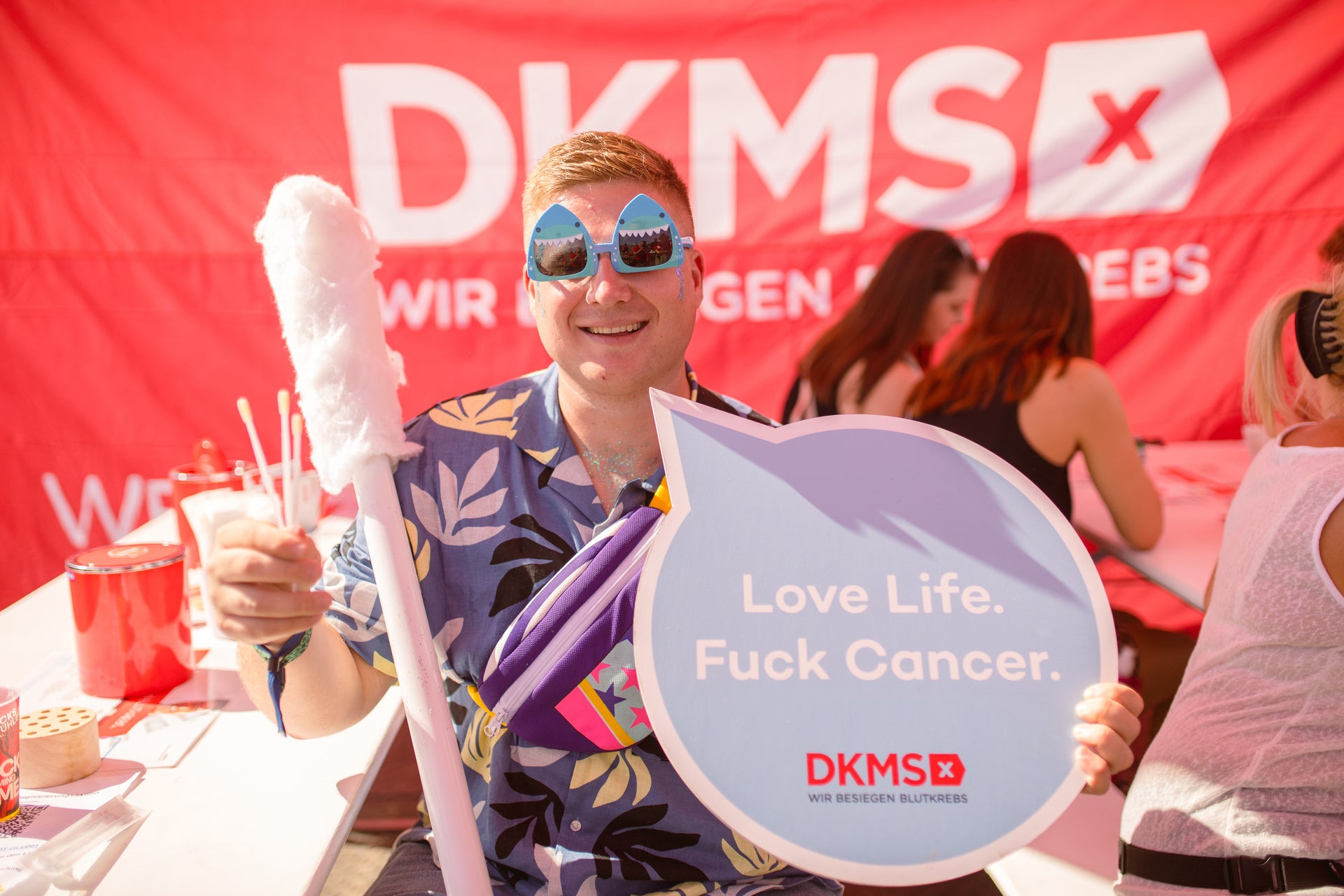 Ein Mann trägt eine Sonnenbrille, hält ein übergroßes Wattestäbchen in der Hand und hält ein Schild mit der Aufschrift: Love Life. Fuck Cancer Ein Mann trägt eine Sonnenbrille, hält ein übergroßes Wattestäbchen in der Hand und hält ein Schild mit der Aufschrift: Love Life. Fuck Cancer