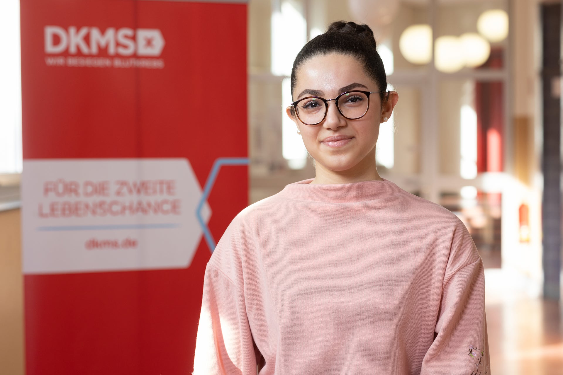 Eine Frau mit Brille und einem rosa Pullover vor einem DKMS Aufsteller Eine Frau mit Brille und einem rosa Pullover vor einem DKMS Aufsteller