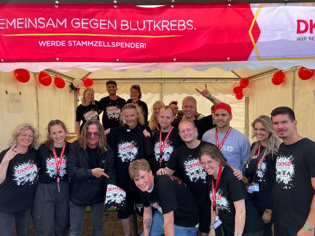 Eine Gruppe von etwa zwanzig Personen steht unter einem roten Banner mit der Aufschrift „Gemeinsam gegen Blutkrebs – Werde Stammzellspender!“ in einem weißen Zelt. Die meisten tragen schwarze DKMS-T-Shirts mit weißem Splash-Design und rote Schlüsselbänder mit Ausweisen. Im Hintergrund hängen rote und weiße Luftballons. Die Gruppe lächelt in die Kamera, einige zeigen Handzeichen. Eine Gruppe von etwa zwanzig Personen steht unter einem roten Banner mit der Aufschrift „Gemeinsam gegen Blutkrebs – Werde Stammzellspender!“ in einem weißen Zelt. Die meisten tragen schwarze DKMS-T-Shirts mit weißem Splash-Design und rote Schlüsselbänder mit Ausweisen. Im Hintergrund hängen rote und weiße Luftballons. Die Gruppe lächelt in die Kamera, einige zeigen Handzeichen.