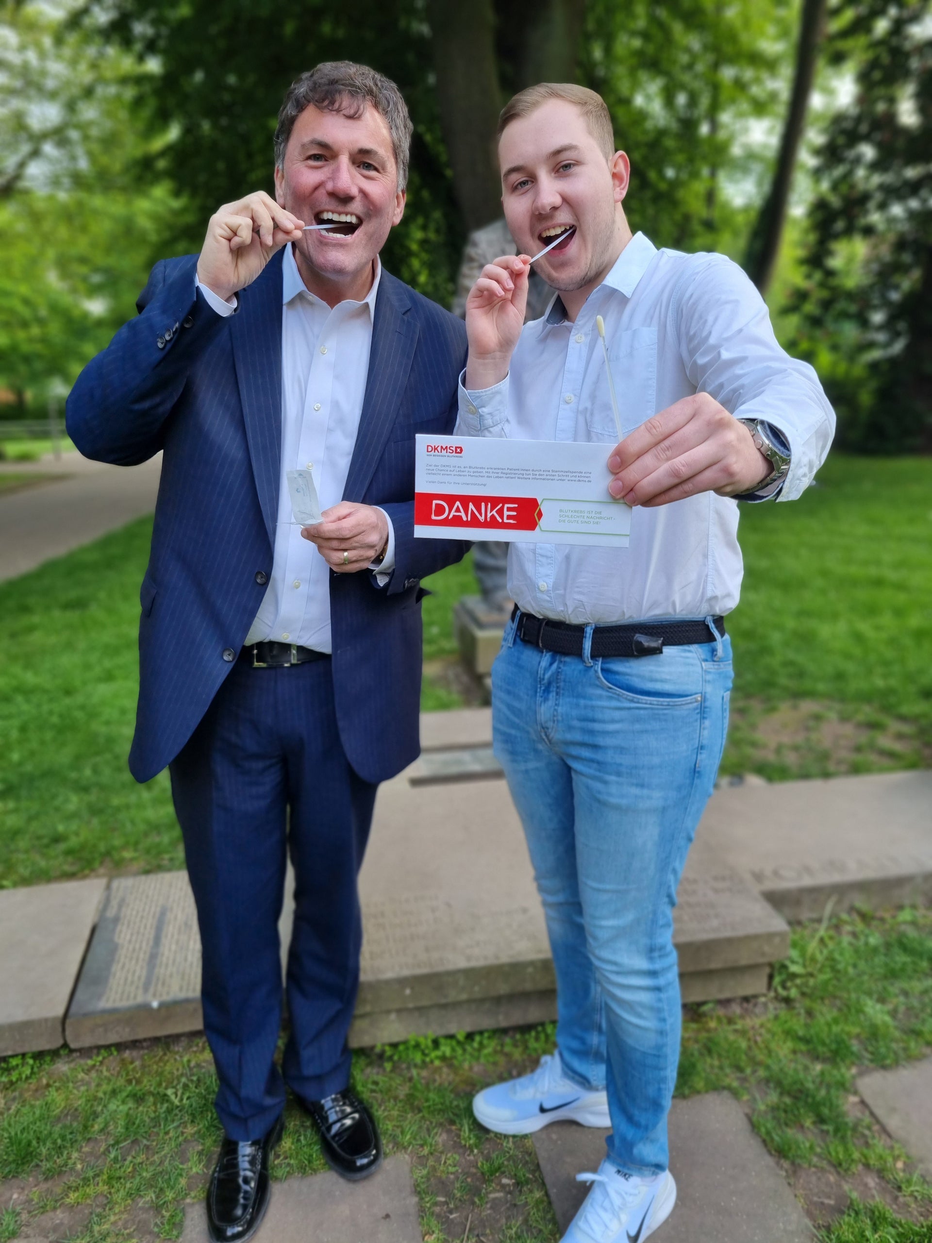 Zwei Männer stehen auf einem Weg im Grünen und führen jeweils ein Wattestäbchen zum Mund. Der Mann rechts hält zusätzlich ein DKMS-Registrierungsset mit dem Schriftzug „DANKE“. Beide tragen helle Hemden, der linke Mann zusätzlich einen dunklen Anzug. Im Hintergrund sind Rasenflächen, Bäume und ein Weg zu sehen. Zwei Männer stehen auf einem Weg im Grünen und führen jeweils ein Wattestäbchen zum Mund. Der Mann rechts hält zusätzlich ein DKMS-Registrierungsset mit dem Schriftzug „DANKE“. Beide tragen helle Hemden, der linke Mann zusätzlich einen dunklen Anzug. Im Hintergrund sind Rasenflächen, Bäume und ein Weg zu sehen.