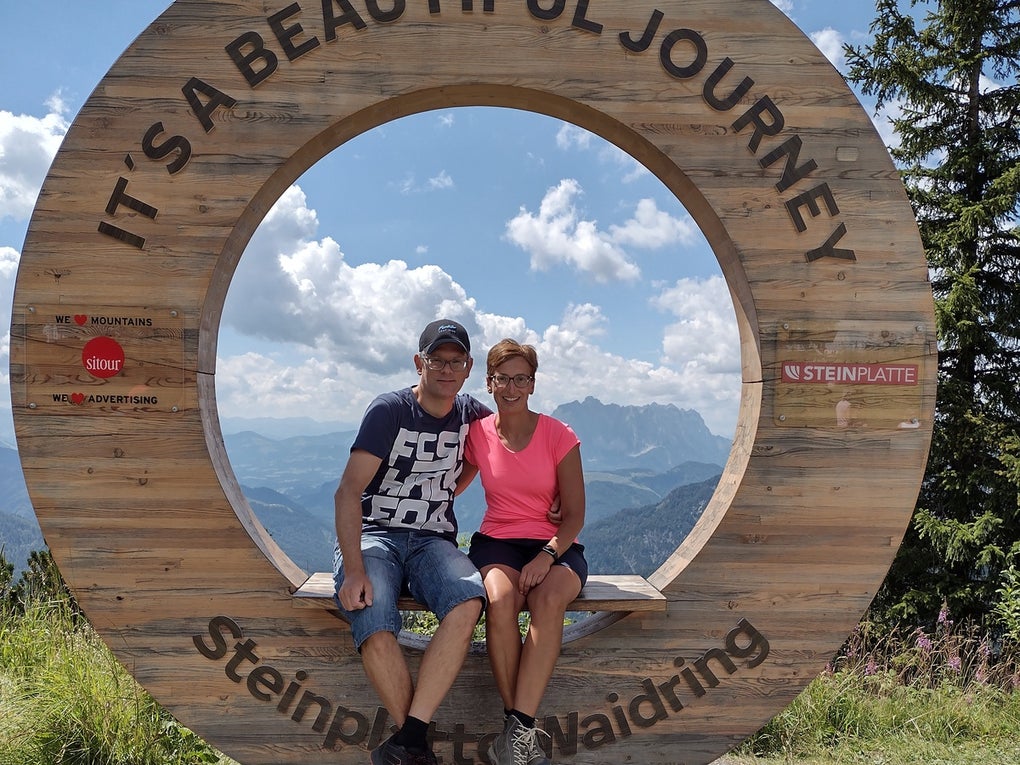 Ein Mann und eine Frau sitzen auf einem hölzernen Rundrahmen mit der Aufschrift „IT'S A BEAUTIFUL JOURNEY – Steinplatte Waidring“. Beide tragen sportliche Kleidung und Wanderschuhe. Im Hintergrund sind Berge, Wolken und blauer Himmel zu sehen. Der Rahmen steht an einem Aussichtspunkt auf einem Wanderweg.
Ein Mann und eine Frau sitzen auf einem hölzernen Rundrahmen mit der Aufschrift „IT'S A BEAUTIFUL JOURNEY – Steinplatte Waidring“. Beide tragen sportliche Kleidung und Wanderschuhe. Im Hintergrund sind Berge, Wolken und blauer Himmel zu sehen. Der Rahmen steht an einem Aussichtspunkt auf einem Wanderweg.