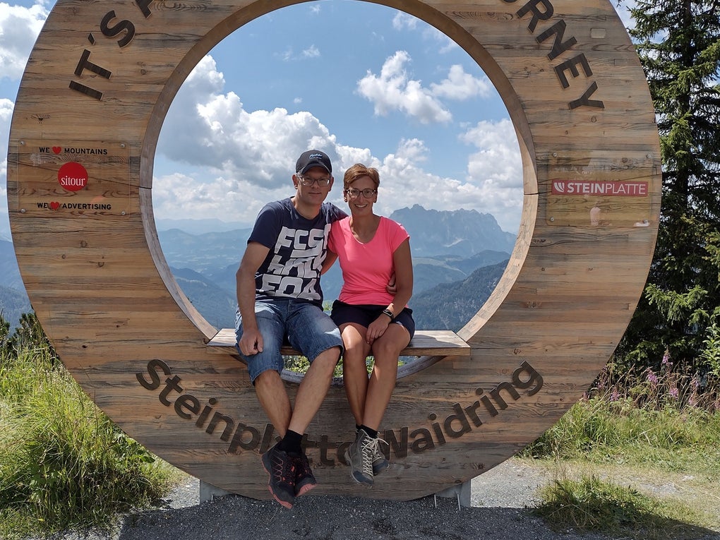 Ein Mann und eine Frau sitzen auf einem hölzernen Rundrahmen mit der Aufschrift „IT'S A BEAUTIFUL JOURNEY – Steinplatte Waidring“. Beide tragen sportliche Kleidung und Wanderschuhe. Im Hintergrund sind Berge, Wolken und blauer Himmel zu sehen. Der Rahmen steht an einem Aussichtspunkt auf einem Wanderweg.
 Ein Mann und eine Frau sitzen auf einem hölzernen Rundrahmen mit der Aufschrift „IT'S A BEAUTIFUL JOURNEY – Steinplatte Waidring“. Beide tragen sportliche Kleidung und Wanderschuhe. Im Hintergrund sind Berge, Wolken und blauer Himmel zu sehen. Der Rahmen steht an einem Aussichtspunkt auf einem Wanderweg.