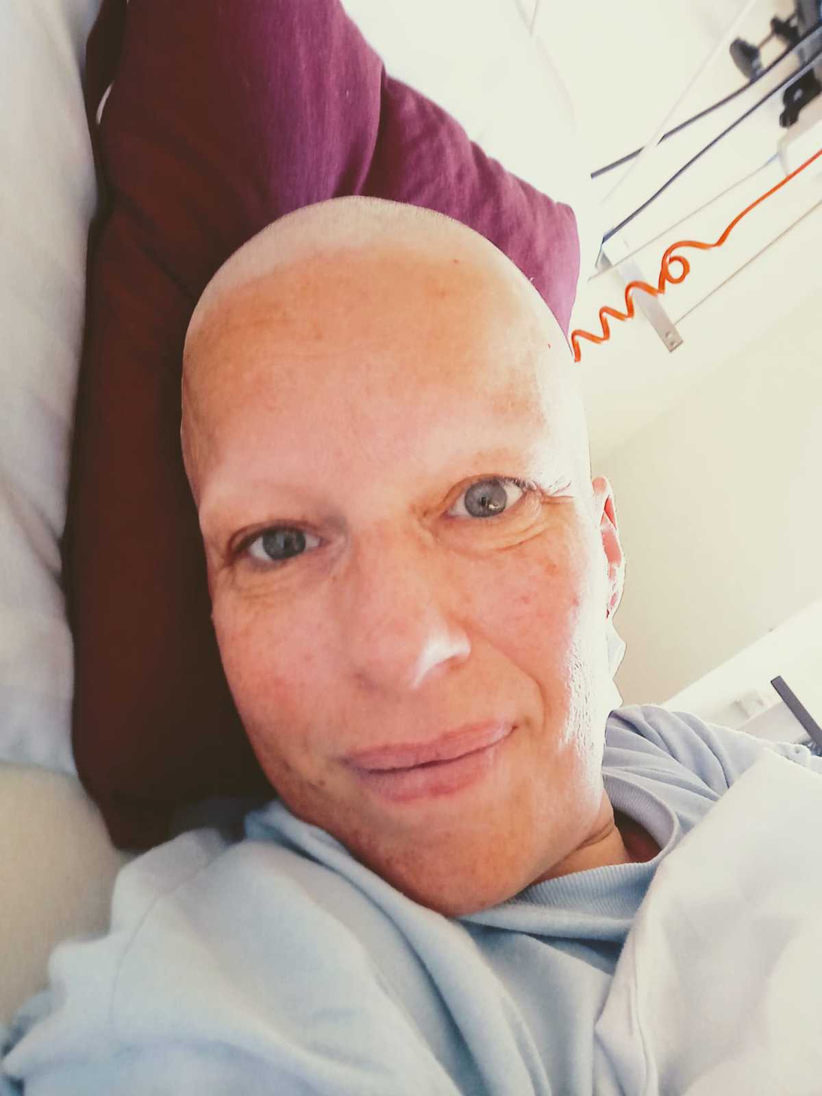 Eine Person mit kahlrasiertem Kopf liegt auf einem Kissen mit dunkelrotem Bezug und blickt direkt in die Kamera. Sie trägt ein hellblaues Oberteil. Im Hintergrund sind Teile eines Krankenhauszimmers sichtbar, darunter ein orangefarbenes gewendeltes Kabel und eine Deckenhalterung.
Eine Person mit kahlrasiertem Kopf liegt auf einem Kissen mit dunkelrotem Bezug und blickt direkt in die Kamera. Sie trägt ein hellblaues Oberteil. Im Hintergrund sind Teile eines Krankenhauszimmers sichtbar, darunter ein orangefarbenes gewendeltes Kabel und eine Deckenhalterung.