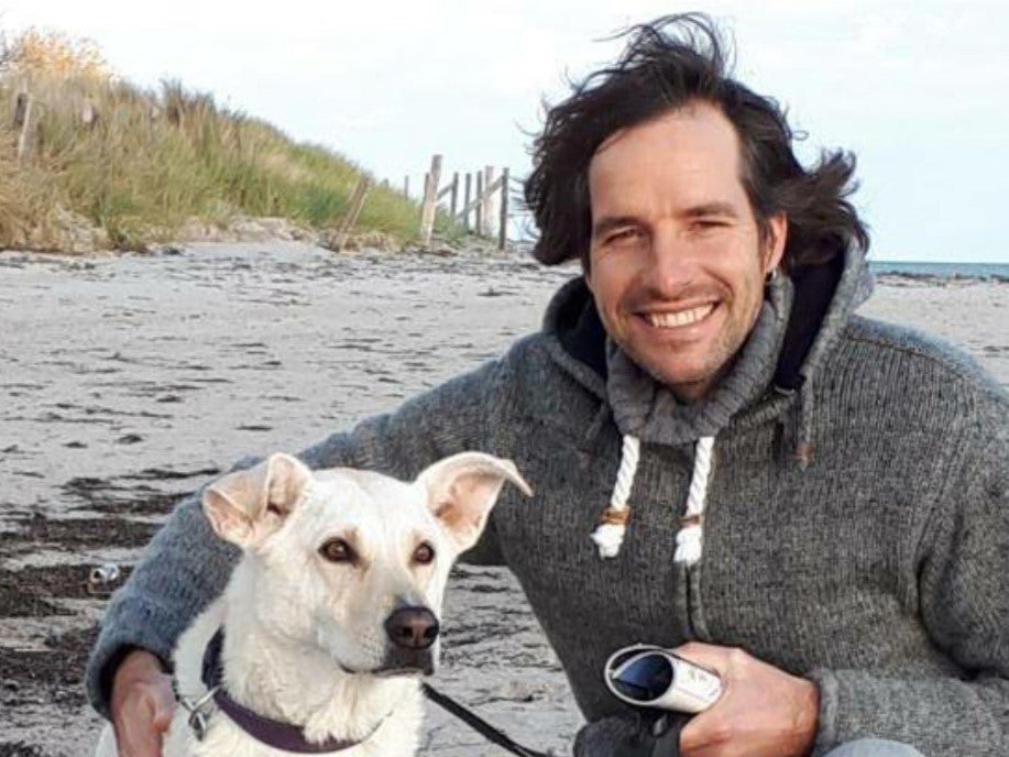 Ein Mann mit dunklem Haar und grauem Kapuzenpullover kniet am Strand neben einem weißen Hund mit Leine. Beide blicken in die Kamera. Der Mann hält eine Kamera in der Hand und lächelt. Im Hintergrund sind Sand, Dünengras und das Meer zu sehen.
Ein Mann mit dunklem Haar und grauem Kapuzenpullover kniet am Strand neben einem weißen Hund mit Leine. Beide blicken in die Kamera. Der Mann hält eine Kamera in der Hand und lächelt. Im Hintergrund sind Sand, Dünengras und das Meer zu sehen.