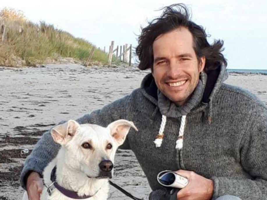 Ein Mann mit dunklem Haar und grauem Kapuzenpullover kniet am Strand neben einem weißen Hund mit Leine. Beide blicken in die Kamera. Der Mann hält eine Kamera in der Hand und lächelt. Im Hintergrund sind Sand, Dünengras und das Meer zu sehen.
Ein Mann mit dunklem Haar und grauem Kapuzenpullover kniet am Strand neben einem weißen Hund mit Leine. Beide blicken in die Kamera. Der Mann hält eine Kamera in der Hand und lächelt. Im Hintergrund sind Sand, Dünengras und das Meer zu sehen.