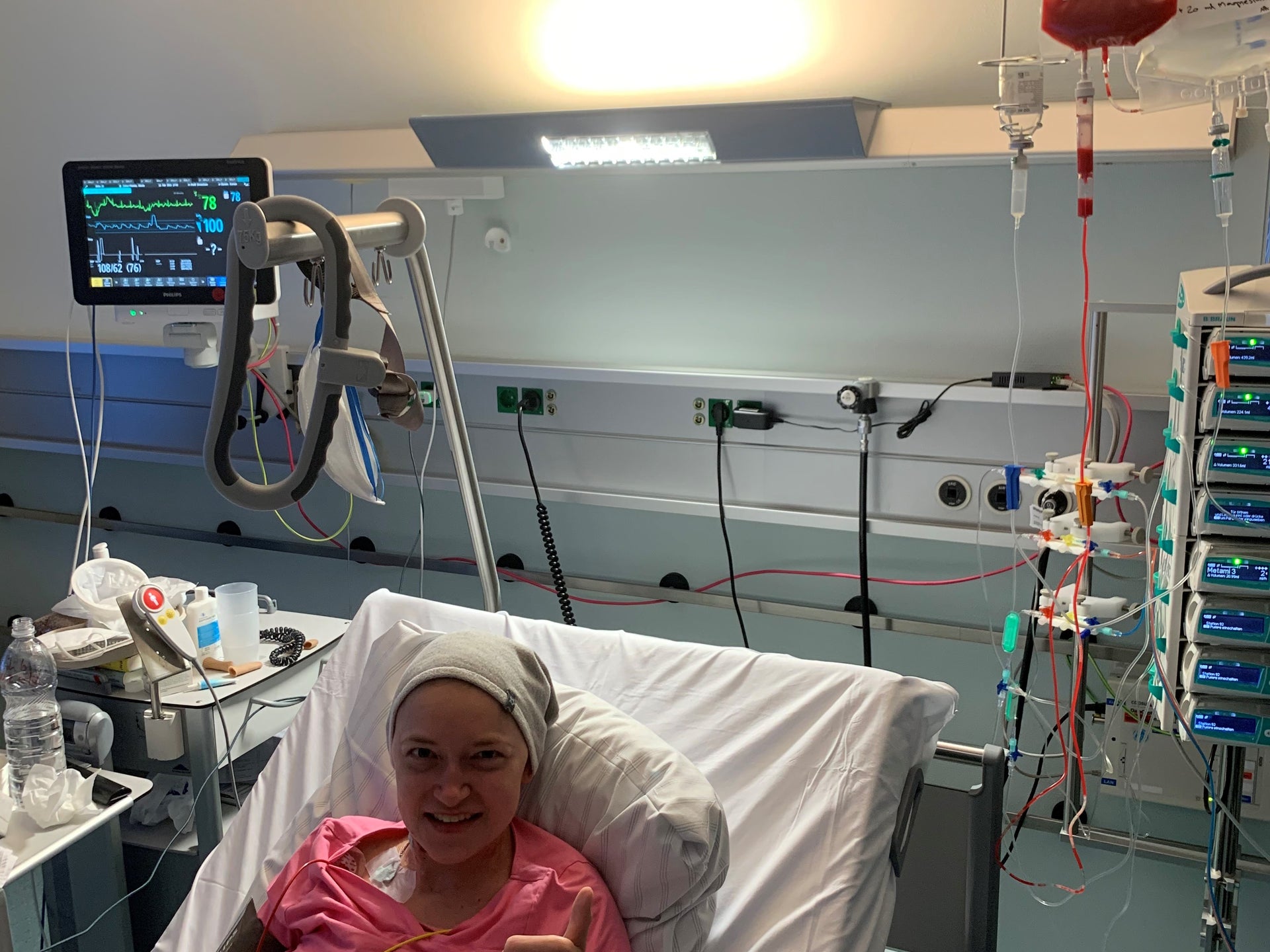 Eine Frau liegt in einem Krankenhausbett und zeigt mit einem Lächeln den Daumen nach oben. Sie trägt ein rosafarbenes Shirt und ein helles Tuch auf dem Kopf. Neben ihr sind medizinische Geräte und Infusionen zu sehen, darunter ein Beutel mit roten Stammzellen. Eine Frau liegt in einem Krankenhausbett und zeigt mit einem Lächeln den Daumen nach oben. Sie trägt ein rosafarbenes Shirt und ein helles Tuch auf dem Kopf. Neben ihr sind medizinische Geräte und Infusionen zu sehen, darunter ein Beutel mit roten Stammzellen.
