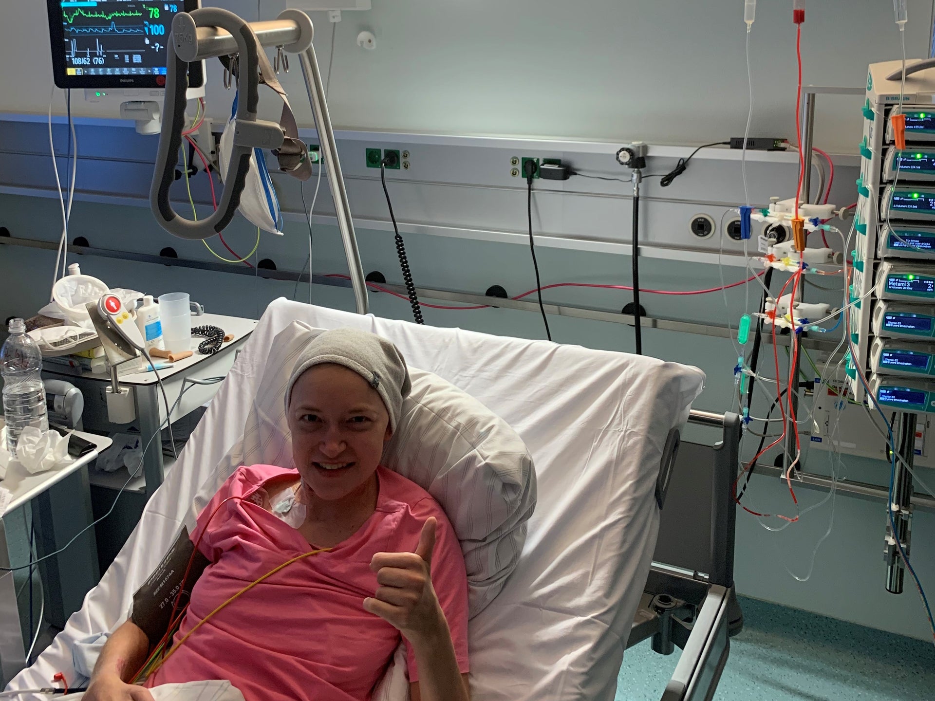 Eine Frau liegt in einem Krankenhausbett und zeigt mit einem Lächeln den Daumen nach oben. Sie trägt ein rosafarbenes Shirt und ein helles Tuch auf dem Kopf. Neben ihr sind medizinische Geräte und Infusionen zu sehen, darunter ein Beutel mit roten Stammzellen. Eine Frau liegt in einem Krankenhausbett und zeigt mit einem Lächeln den Daumen nach oben. Sie trägt ein rosafarbenes Shirt und ein helles Tuch auf dem Kopf. Neben ihr sind medizinische Geräte und Infusionen zu sehen, darunter ein Beutel mit roten Stammzellen.