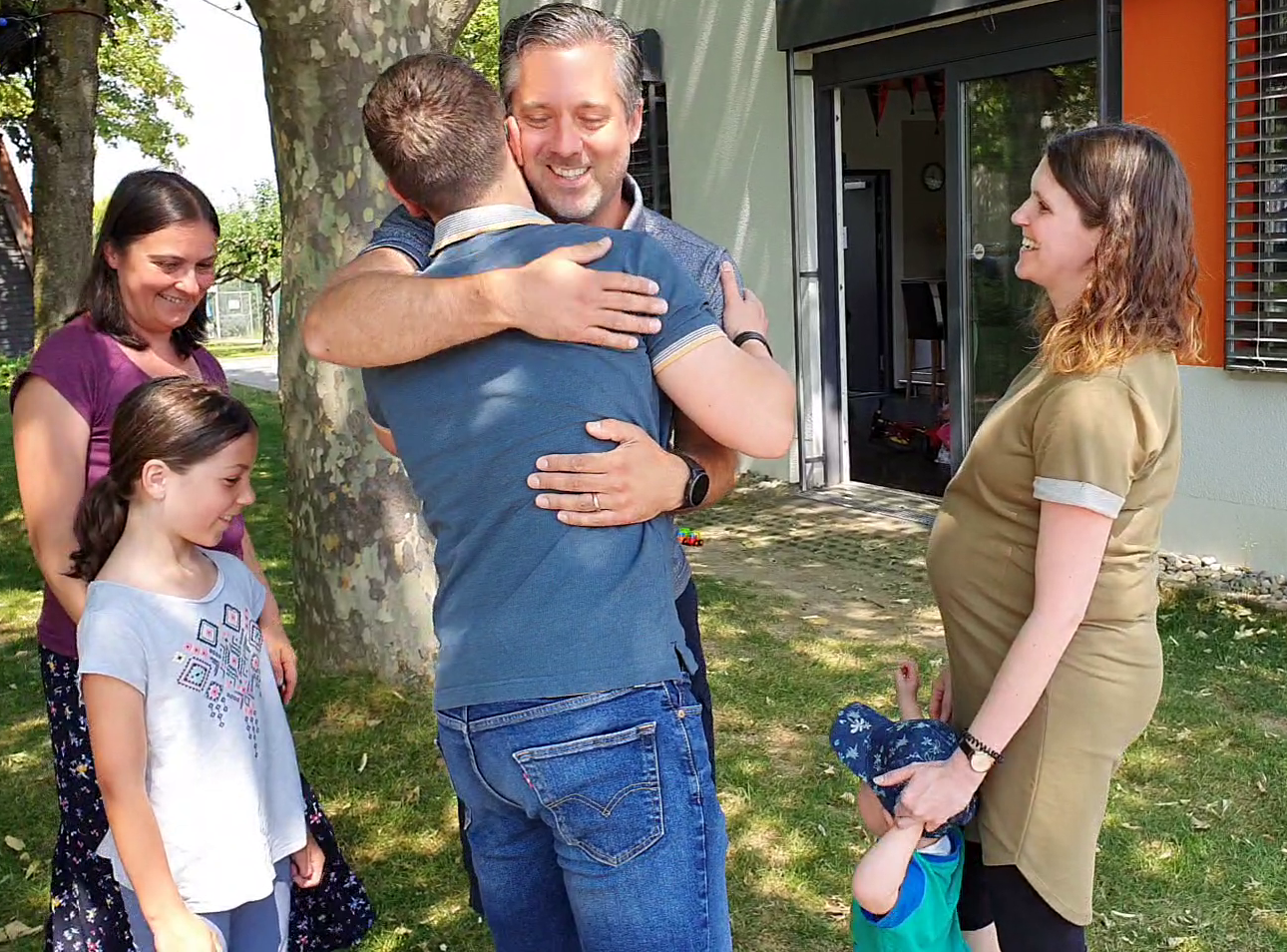 Zwei Männer umarmen sich herzlich im Freien vor einem Gebäude mit orangefarbener Fensterleiste. Um sie herum stehen vier weitere Personen: eine Frau mit einem kleinen Kind an der Hand, eine weitere Frau und ein Mädchen. Im Hintergrund sind ein Baum, Rasenfläche und Gebäudeteile mit Fensterläden zu sehen.
Zwei Männer umarmen sich herzlich im Freien vor einem Gebäude mit orangefarbener Fensterleiste. Um sie herum stehen vier weitere Personen: eine Frau mit einem kleinen Kind an der Hand, eine weitere Frau und ein Mädchen. Im Hintergrund sind ein Baum, Rasenfläche und Gebäudeteile mit Fensterläden zu sehen.