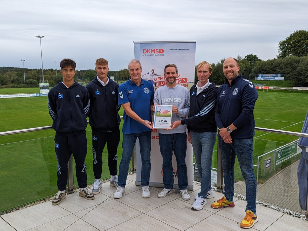 Sechs Personen stehen auf einer Terrasse vor einem Sportplatz. Vier junge Männer tragen Sportkleidung mit dem Logo eines Fußballvereins. Eine Frau in der Mitte hält zusammen mit einem Mann ein DKMS-Zertifikat. Im Hintergrund ist ein DKMS-Rollup mit dem Text „Vereint gegen Blutkrebs“ zu sehen. Es ist bewölkt, die Umgebung ist grün mit Bäumen und Rasenfläche. Sechs Personen stehen auf einer Terrasse vor einem Sportplatz. Vier junge Männer tragen Sportkleidung mit dem Logo eines Fußballvereins. Eine Frau in der Mitte hält zusammen mit einem Mann ein DKMS-Zertifikat. Im Hintergrund ist ein DKMS-Rollup mit dem Text „Vereint gegen Blutkrebs“ zu sehen. Es ist bewölkt, die Umgebung ist grün mit Bäumen und Rasenfläche.