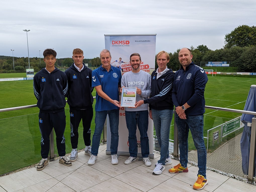 Sechs Personen stehen auf einer Terrasse vor einem Sportplatz. Vier junge Männer tragen Sportkleidung mit dem Logo eines Fußballvereins. Eine Frau in der Mitte hält zusammen mit einem Mann ein DKMS-Zertifikat. Im Hintergrund ist ein DKMS-Rollup mit dem Text „Vereint gegen Blutkrebs“ zu sehen. Es ist bewölkt, die Umgebung ist grün mit Bäumen und Rasenfläche. Sechs Personen stehen auf einer Terrasse vor einem Sportplatz. Vier junge Männer tragen Sportkleidung mit dem Logo eines Fußballvereins. Eine Frau in der Mitte hält zusammen mit einem Mann ein DKMS-Zertifikat. Im Hintergrund ist ein DKMS-Rollup mit dem Text „Vereint gegen Blutkrebs“ zu sehen. Es ist bewölkt, die Umgebung ist grün mit Bäumen und Rasenfläche.