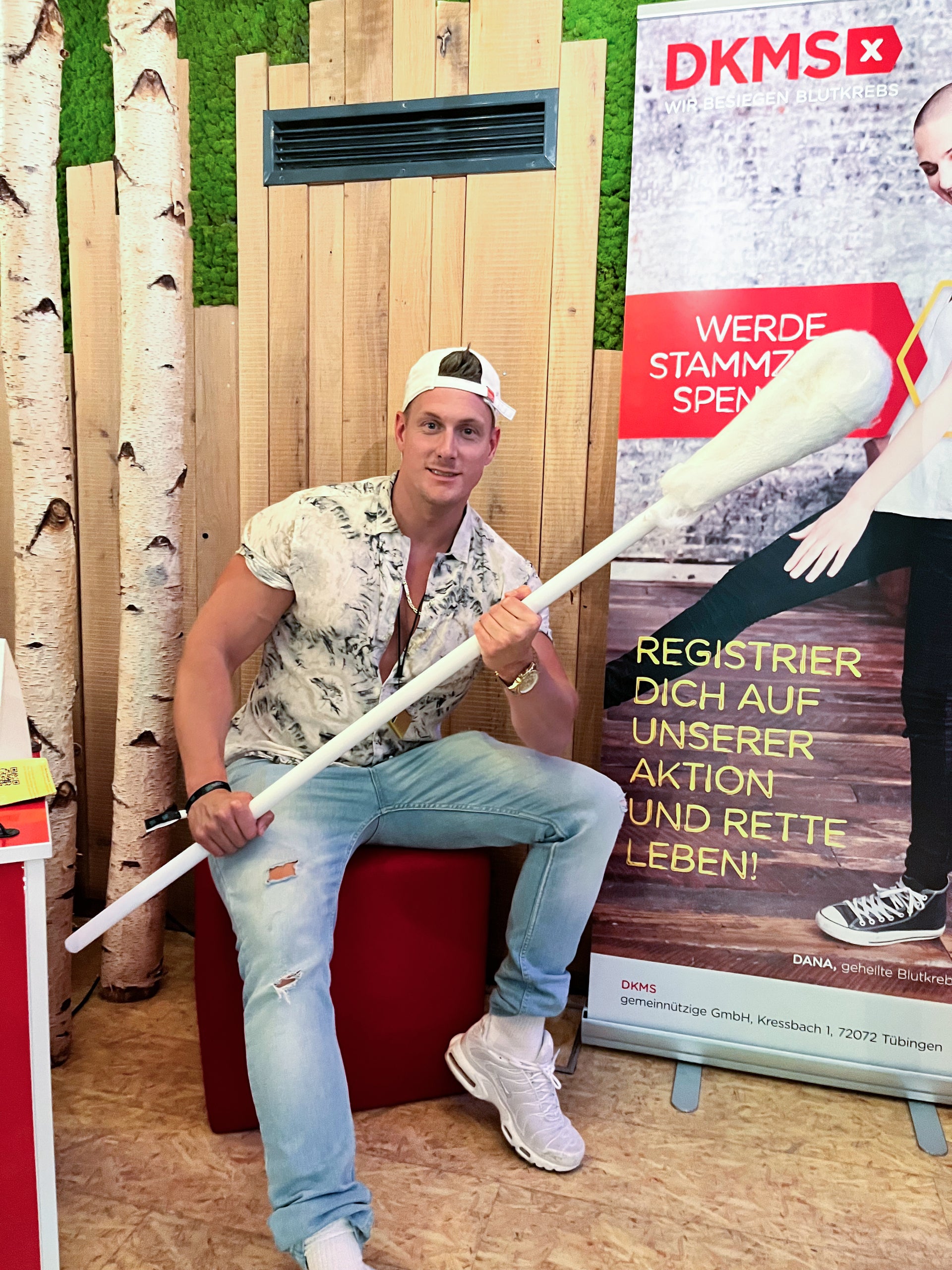 Ein Mann mit Basecap sitzt auf einem roten Hocker und hält ein übergroßes Wattestäbchen in den Händen. Er trägt ein gemustertes Hemd, eine helle Jeans und weiße Turnschuhe. Hinter ihm steht ein DKMS-Rollup mit der Aufschrift „Werde Stammzellspender!“ sowie „Registrier dich auf unserer Aktion und rette Leben!“. Links im Hintergrund sind Holzpaneele und Dekobirkenstämme zu sehen.
Ein Mann mit Basecap sitzt auf einem roten Hocker und hält ein übergroßes Wattestäbchen in den Händen. Er trägt ein gemustertes Hemd, eine helle Jeans und weiße Turnschuhe. Hinter ihm steht ein DKMS-Rollup mit der Aufschrift „Werde Stammzellspender!“ sowie „Registrier dich auf unserer Aktion und rette Leben!“. Links im Hintergrund sind Holzpaneele und Dekobirkenstämme zu sehen.
