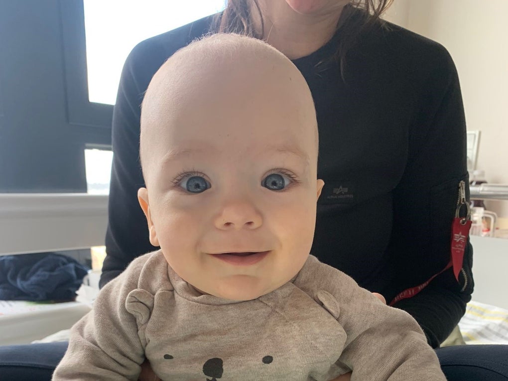 Ein Baby mit großen blauen Augen und einem beigefarbenen Pullover mit Bärengesicht sitzt auf dem Schoß einer Frau. Das Baby schaut lächelnd in die Kamera. Im Hintergrund ist ein Fenster und ein helles Zimmer zu sehen. Ein Baby mit großen blauen Augen und einem beigefarbenen Pullover mit Bärengesicht sitzt auf dem Schoß einer Frau. Das Baby schaut lächelnd in die Kamera. Im Hintergrund ist ein Fenster und ein helles Zimmer zu sehen.