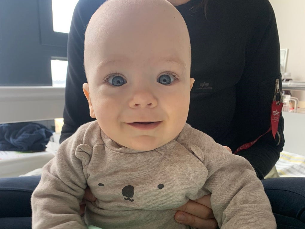 Ein Baby mit großen blauen Augen und einem beigefarbenen Pullover mit Bärengesicht sitzt auf dem Schoß einer Frau. Das Baby schaut lächelnd in die Kamera. Im Hintergrund ist ein Fenster und ein helles Zimmer zu sehen. Ein Baby mit großen blauen Augen und einem beigefarbenen Pullover mit Bärengesicht sitzt auf dem Schoß einer Frau. Das Baby schaut lächelnd in die Kamera. Im Hintergrund ist ein Fenster und ein helles Zimmer zu sehen.