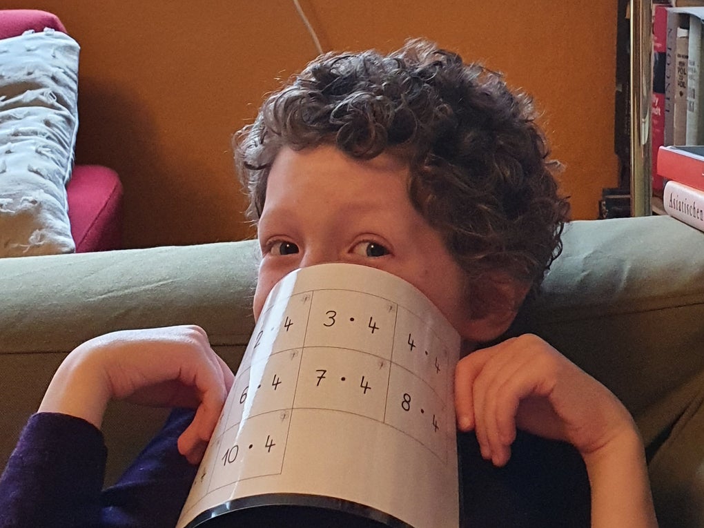 Ein Kind mit lockigen Haaren sitzt auf einem Sofa und hält sich ein Blatt Papier mit Rechenaufgaben spielerisch vor den Mund. Das Kind blickt mit einem verschmitzten Ausdruck zur Seite. Im Hintergrund sind ein rotes Kissen, Bücherregale und ein orangefarbener Wandbereich zu sehen. Ein Kind mit lockigen Haaren sitzt auf einem Sofa und hält sich ein Blatt Papier mit Rechenaufgaben spielerisch vor den Mund. Das Kind blickt mit einem verschmitzten Ausdruck zur Seite. Im Hintergrund sind ein rotes Kissen, Bücherregale und ein orangefarbener Wandbereich zu sehen.