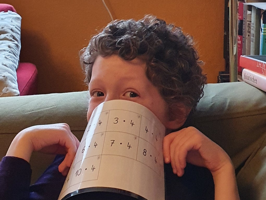 Ein Kind mit lockigen Haaren sitzt auf einem Sofa und hält sich ein Blatt Papier mit Rechenaufgaben spielerisch vor den Mund. Das Kind blickt mit einem verschmitzten Ausdruck zur Seite. Im Hintergrund sind ein rotes Kissen, Bücherregale und ein orangefarbener Wandbereich zu sehen. Ein Kind mit lockigen Haaren sitzt auf einem Sofa und hält sich ein Blatt Papier mit Rechenaufgaben spielerisch vor den Mund. Das Kind blickt mit einem verschmitzten Ausdruck zur Seite. Im Hintergrund sind ein rotes Kissen, Bücherregale und ein orangefarbener Wandbereich zu sehen.