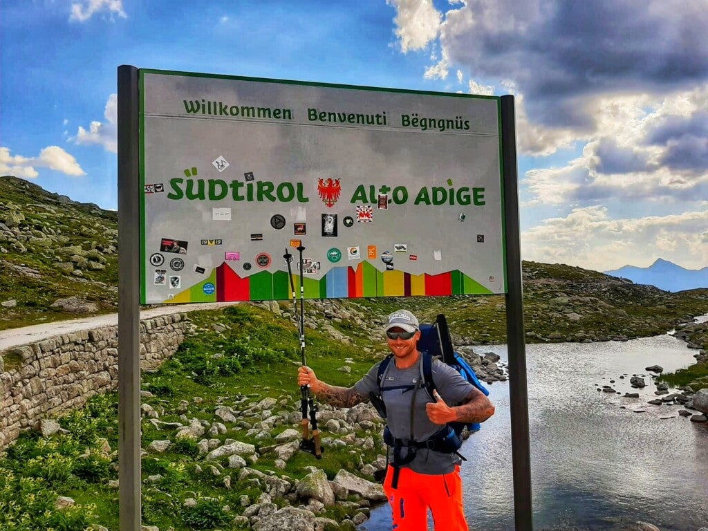 Ein Mann steht vor einem Schild auf dem in verschiedenen Sprachen Wilkommen steht. Darunter steht Südtirol Alto Adige Ein Mann steht vor einem Schild auf dem in verschiedenen Sprachen Wilkommen steht. Darunter steht Südtirol Alto Adige