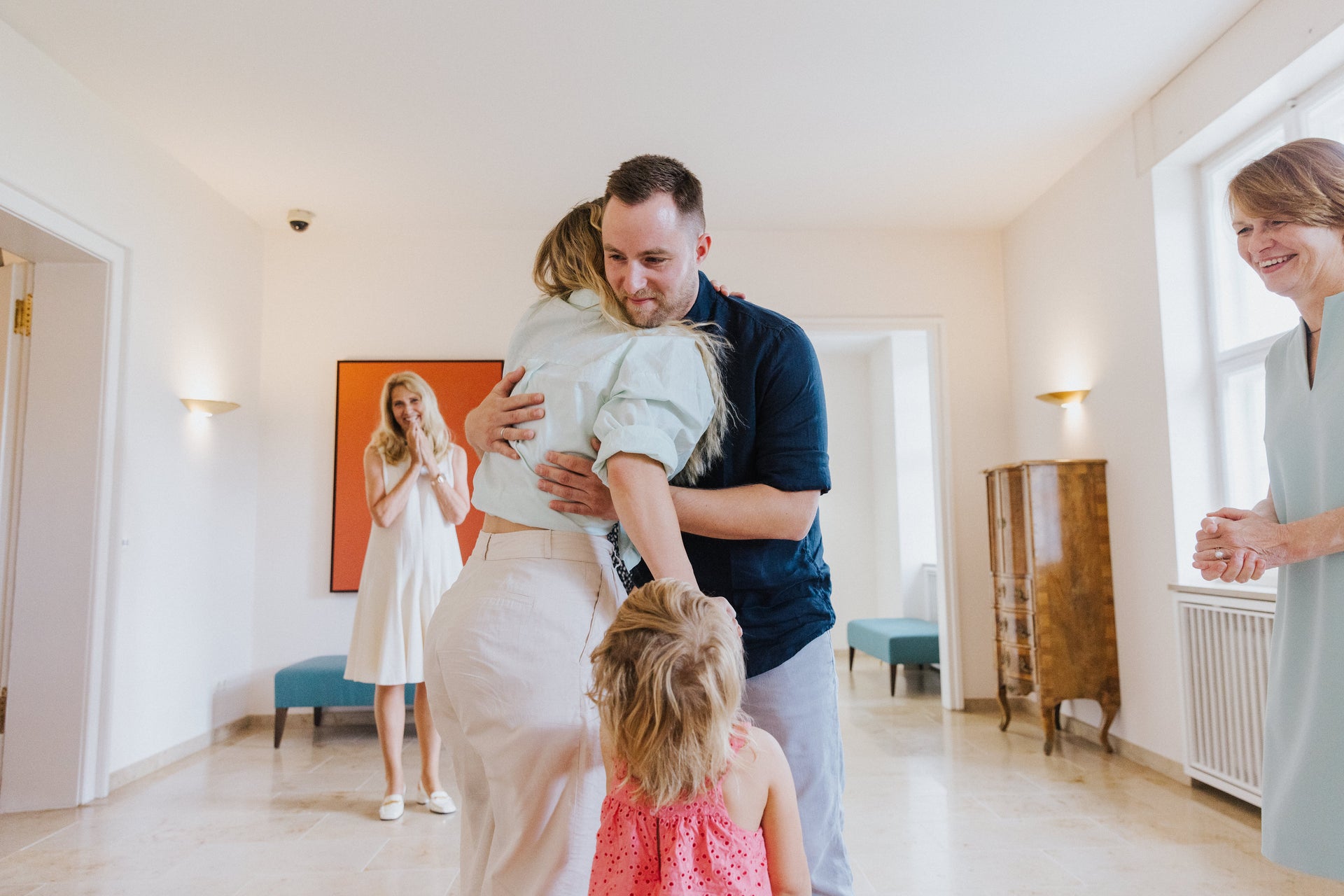 Ein Mann und eine Frau umarmen sich in einem hellen Raum, ein kleines Mädchen steht vor ihnen und berührt sie. Im Hintergrund stehen drei Frauen, zwei davon lächeln, eine klatscht in die Hände. Der Raum ist mit hellen Wänden, großen Fenstern, einem Holzschrank und einem orangefarbenen Bild gestaltet. Ein Mann und eine Frau umarmen sich in einem hellen Raum, ein kleines Mädchen steht vor ihnen und berührt sie. Im Hintergrund stehen drei Frauen, zwei davon lächeln, eine klatscht in die Hände. Der Raum ist mit hellen Wänden, großen Fenstern, einem Holzschrank und einem orangefarbenen Bild gestaltet.