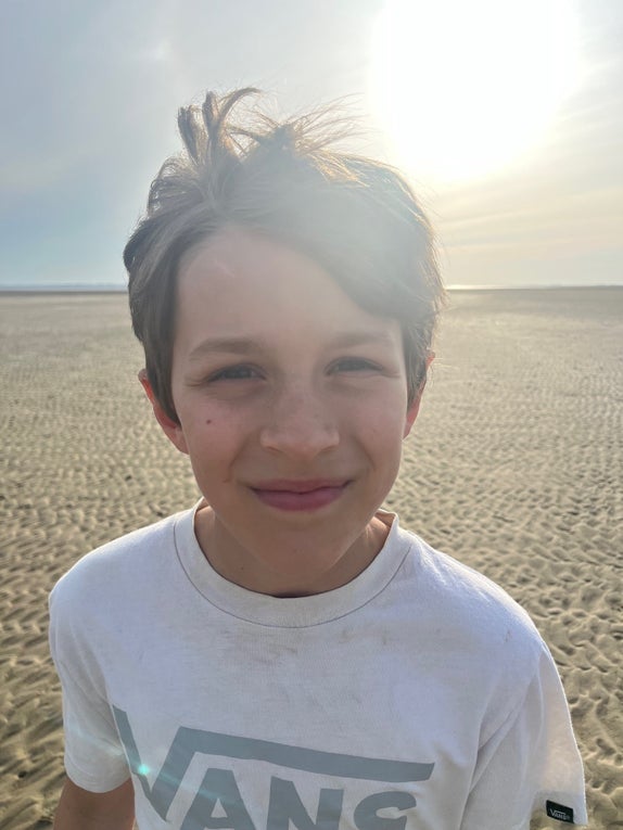 Ein Junge mit braunen Haaren und weißem T-Shirt lächelt in die Kamera. Hinter ihm ist die Sonne und ein Strand zu sehen. Ein Junge mit braunen Haaren und weißem T-Shirt lächelt in die Kamera. Hinter ihm ist die Sonne und ein Strand zu sehen.