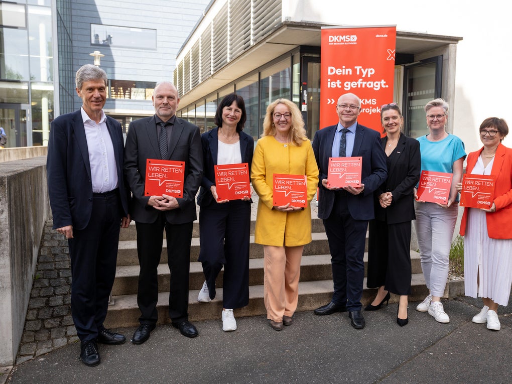 Acht Personen stehen vor einem modernen Gebäude und halten rote DKMS-Schilder mit der Aufschrift „Wir retten Leben“. Im Hintergrund steht ein DKMS-Aufsteller mit der Botschaft „Dein Typ ist gefragt“. Die Gruppe wirkt fröhlich und engagiert. Acht Personen stehen vor einem modernen Gebäude und halten rote DKMS-Schilder mit der Aufschrift „Wir retten Leben“. Im Hintergrund steht ein DKMS-Aufsteller mit der Botschaft „Dein Typ ist gefragt“. Die Gruppe wirkt fröhlich und engagiert.