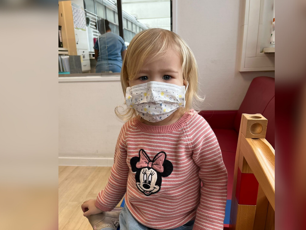 Ein Mädchen sitzt mit Mundschutz und einem Minnie-Maus-Oberteil auf einer Spielmatte. Ein Mädchen sitzt mit Mundschutz und einem Minnie-Maus-Oberteil auf einer Spielmatte.