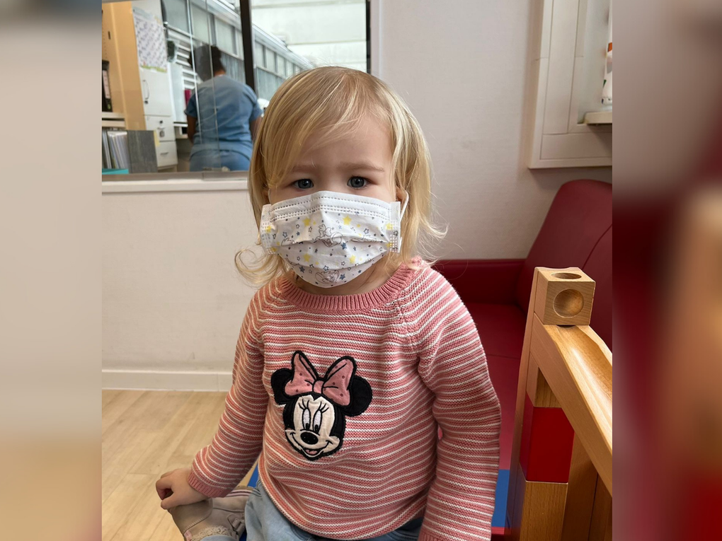 Ein Mädchen sitzt mit Mundschutz und einem Minnie-Maus-Oberteil auf einer Spielmatte. Ein Mädchen sitzt mit Mundschutz und einem Minnie-Maus-Oberteil auf einer Spielmatte.