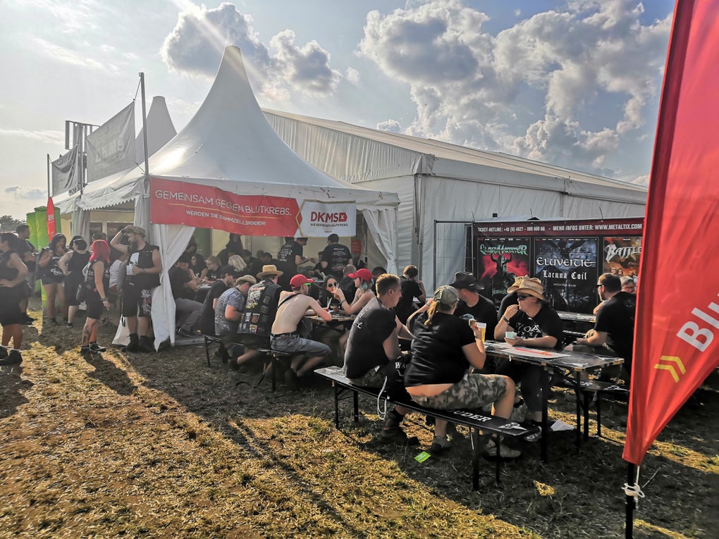DKMS auf dem Wacken Open Air Festival, im Hintergrund der Stand, im Vordergrund viele Leute auf Bierbänken DKMS auf dem Wacken Open Air Festival, im Hintergrund der Stand, im Vordergrund viele Leute auf Bierbänken