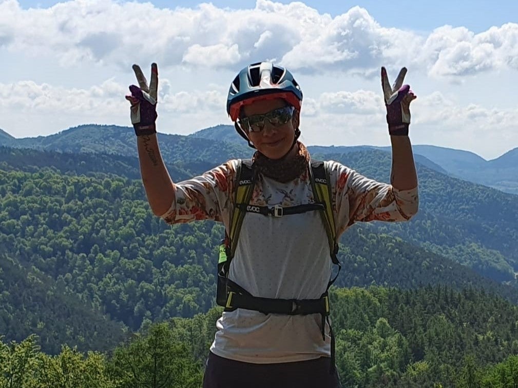 Eine Frau steht mit Fahrradhelm und Rennbrille auf einem Berg und hält Peace-Zeichen in die Luft. Eine Frau steht mit Fahrradhelm und Rennbrille auf einem Berg und hält Peace-Zeichen in die Luft.
