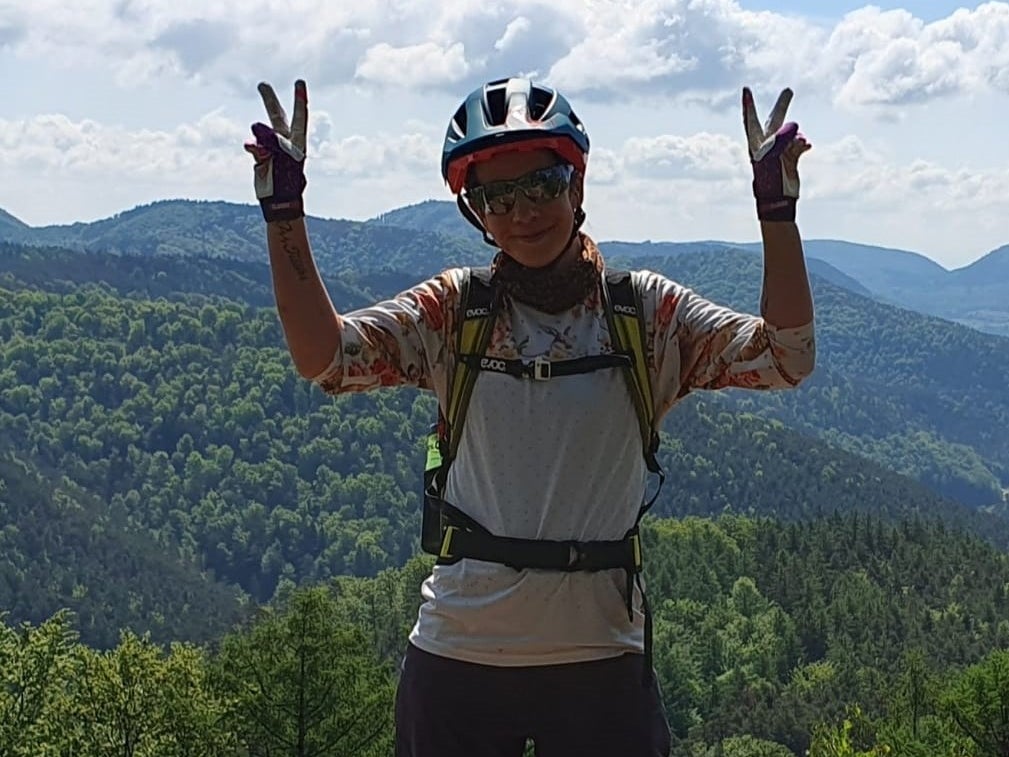 Eine Frau steht mit Fahrradhelm und Rennbrille auf einem Berg und hält Peace-Zeichen in die Luft. Eine Frau steht mit Fahrradhelm und Rennbrille auf einem Berg und hält Peace-Zeichen in die Luft.