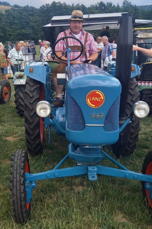 Ein Mann in rotkariertem Hemd und Lederhosen sitzt auf einem blauen Traktor. Ein Mann in rotkariertem Hemd und Lederhosen sitzt auf einem blauen Traktor.