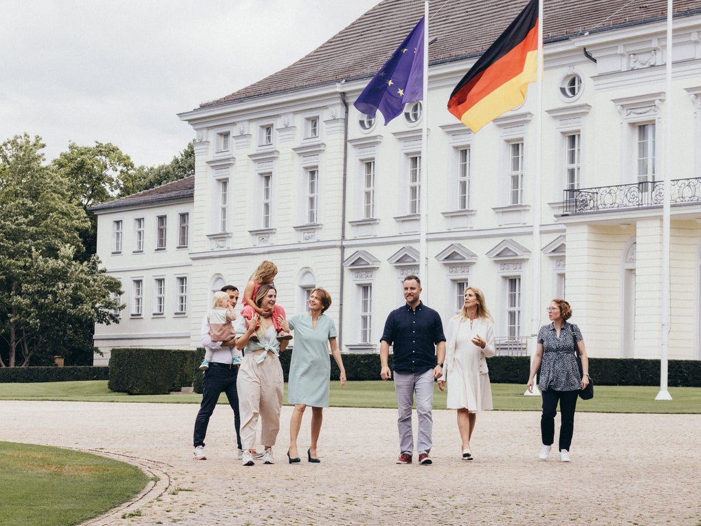 Sechs Erwachsene und zwei Kinder gehen gemeinsam über einen gepflasterten Platz vor einem weißen Gebäude mit Flaggen von Deutschland und der Europäischen Union. Zwei Personen tragen jeweils ein Kind auf dem Arm. Die Gruppe bewegt sich nebeneinander in entspannter Atmosphäre. Im Hintergrund sind Fenster, Balkone und Hecken zu sehen. Sechs Erwachsene und zwei Kinder gehen gemeinsam über einen gepflasterten Platz vor einem weißen Gebäude mit Flaggen von Deutschland und der Europäischen Union. Zwei Personen tragen jeweils ein Kind auf dem Arm. Die Gruppe bewegt sich nebeneinander in entspannter Atmosphäre. Im Hintergrund sind Fenster, Balkone und Hecken zu sehen.