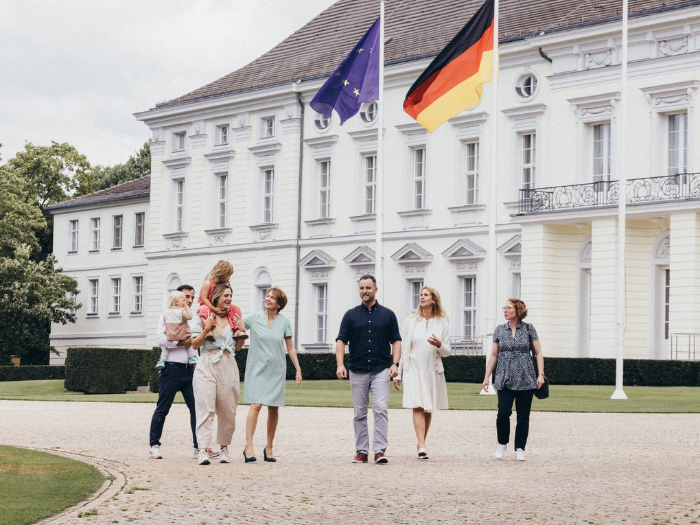 Sechs Erwachsene und zwei Kinder gehen gemeinsam über einen gepflasterten Platz vor einem weißen Gebäude mit Flaggen von Deutschland und der Europäischen Union. Zwei Personen tragen jeweils ein Kind auf dem Arm. Die Gruppe bewegt sich nebeneinander in entspannter Atmosphäre. Im Hintergrund sind Fenster, Balkone und Hecken zu sehen. Sechs Erwachsene und zwei Kinder gehen gemeinsam über einen gepflasterten Platz vor einem weißen Gebäude mit Flaggen von Deutschland und der Europäischen Union. Zwei Personen tragen jeweils ein Kind auf dem Arm. Die Gruppe bewegt sich nebeneinander in entspannter Atmosphäre. Im Hintergrund sind Fenster, Balkone und Hecken zu sehen.