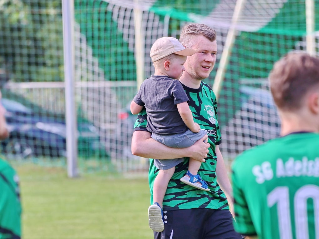ein Mann steht auf einem Rasen, hinter ihm ein Fußballtor und er hält ein Kind auf dem Arm ein Mann steht auf einem Rasen, hinter ihm ein Fußballtor und er hält ein Kind auf dem Arm